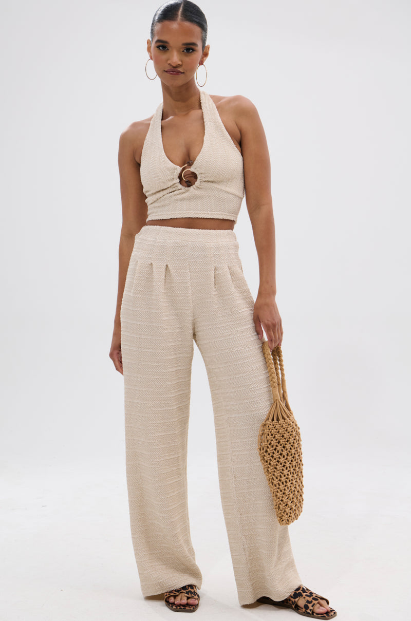 A woman stands confidently against a plain backdrop, wearing the HOLLYWOOD SLINKY TEXTURED PALAZZO PANT in a wide-leg silhouette with a beige halter crop top, hoop earrings, sandals, and a woven bag.
