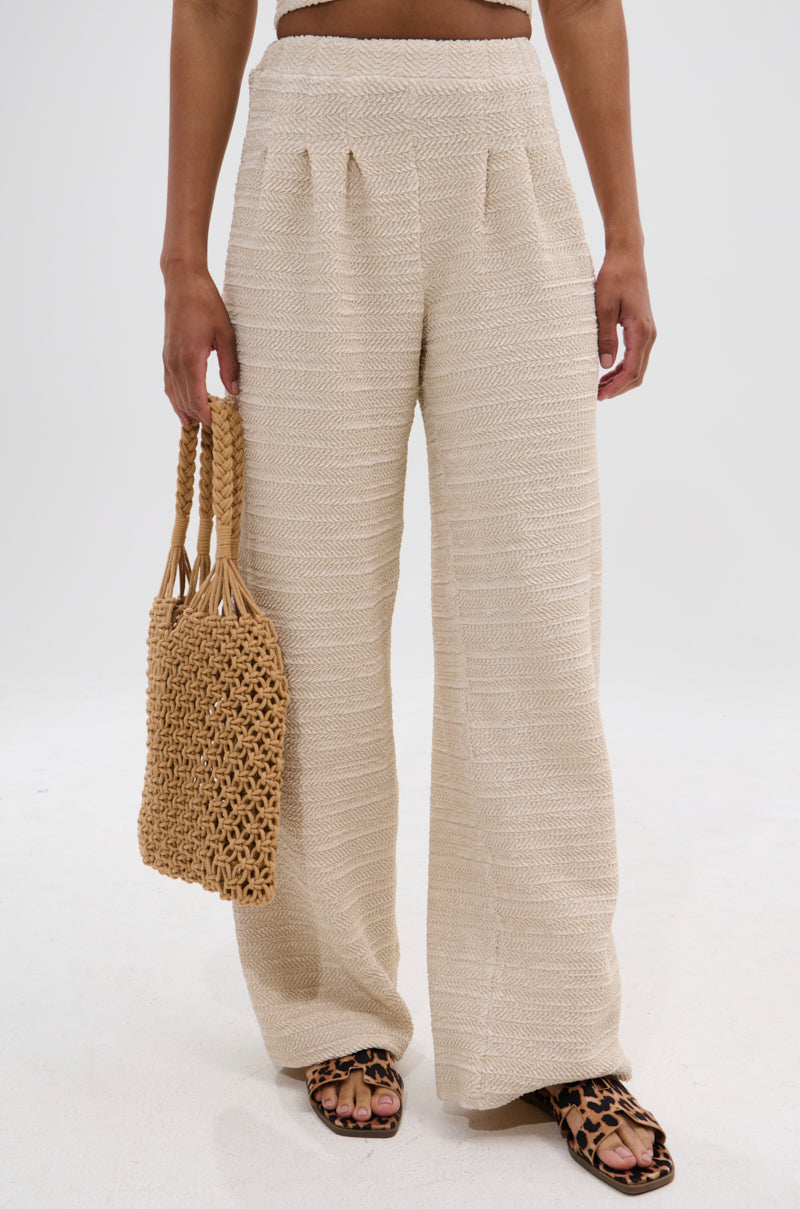 Wearing the HOLLYWOOD SLINKY TEXTURED PALAZZO PANT and leopard print sandals, a person holds a woven straw bag while standing against a plain white background.