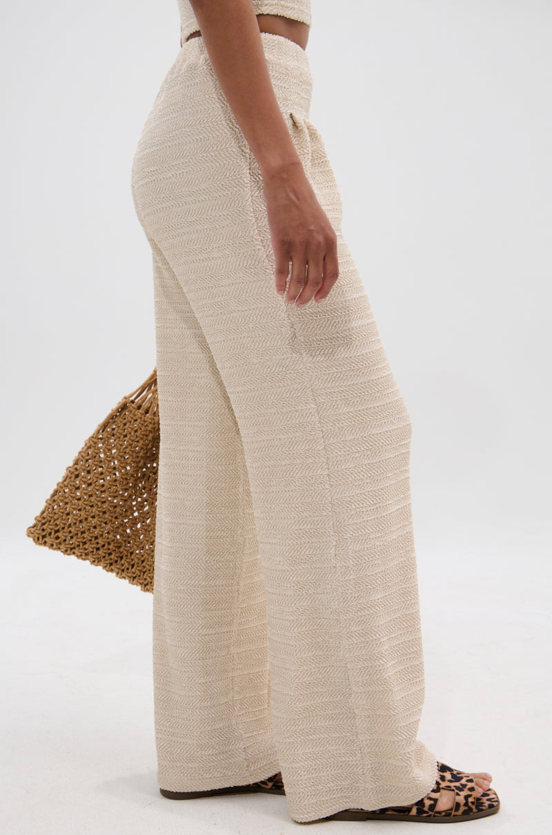 Wearing the HOLLYWOOD SLINKY TEXTURED PALAZZO PANT, a person stands sideways with a woven tan bag and animal print sandals—creating an effortlessly chic look against a plain white background.