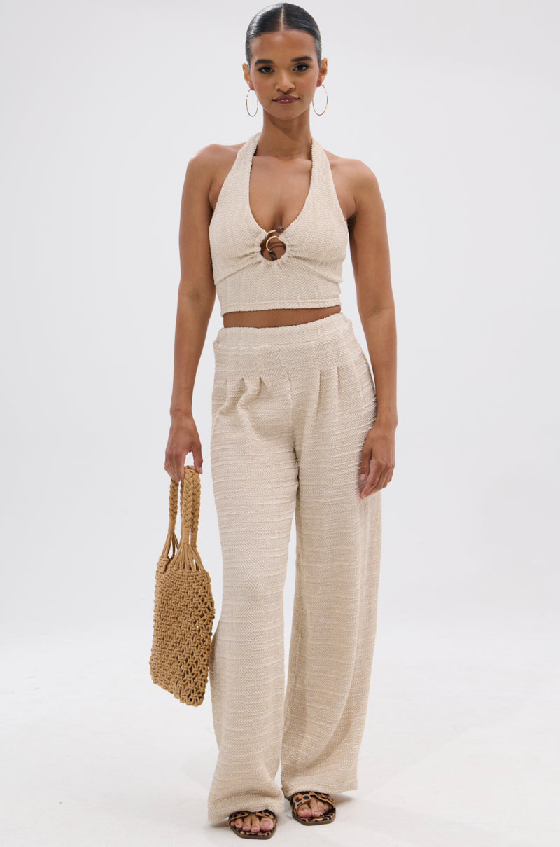 A woman stands against a white background wearing HOLLYWOOD SLINKY TEXTURED PALAZZO PANT, a beige halter crop top with ring detail, hoop earrings, sandals, and holding a woven handbag.
