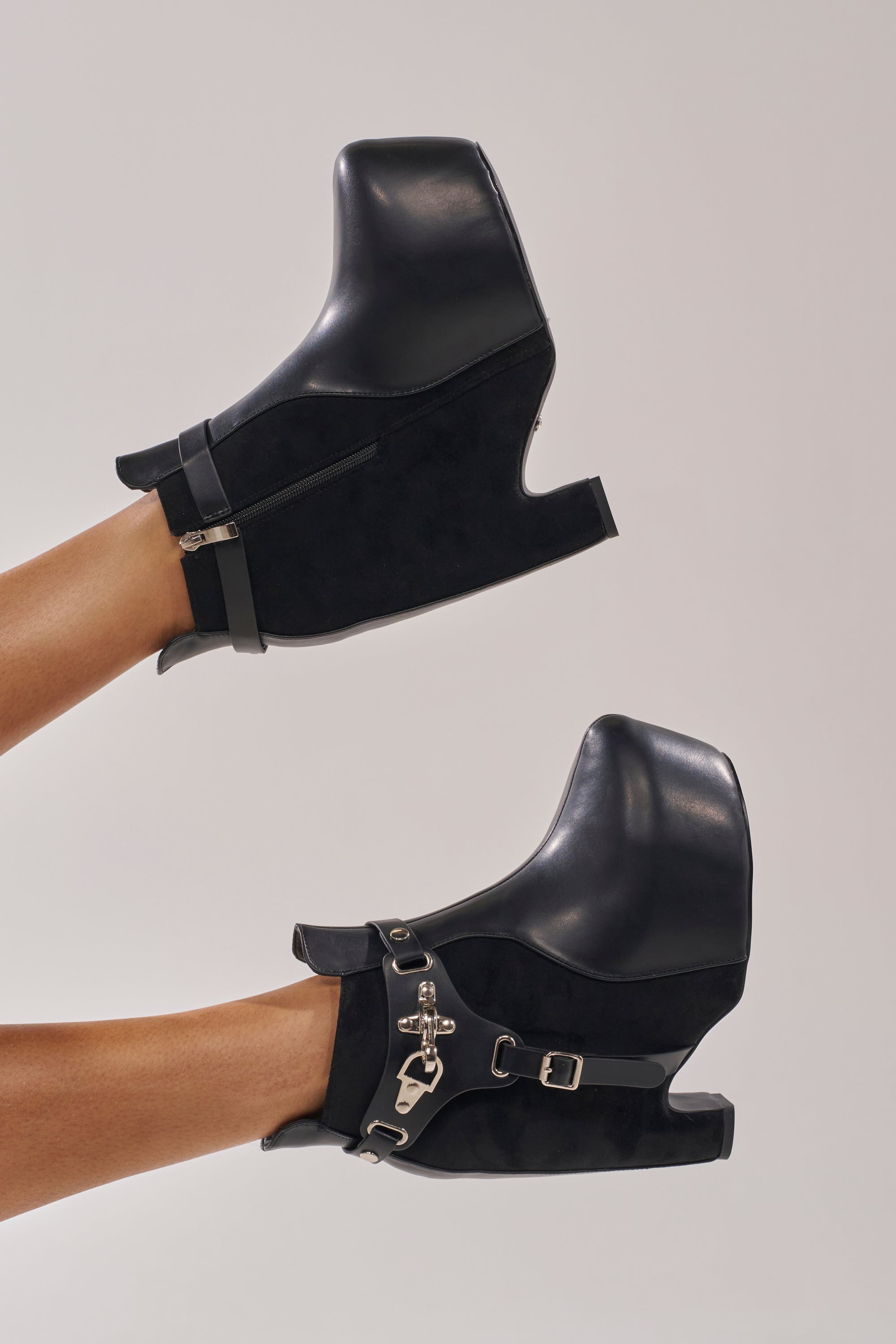 Feet in AZALEA WANG ABYSS BLACK WEDGE PLATFORM BOOTIE—black faux leather booties with chunky curved wedge heels, silver zipper and buckle details—raised against a plain light background. 0
