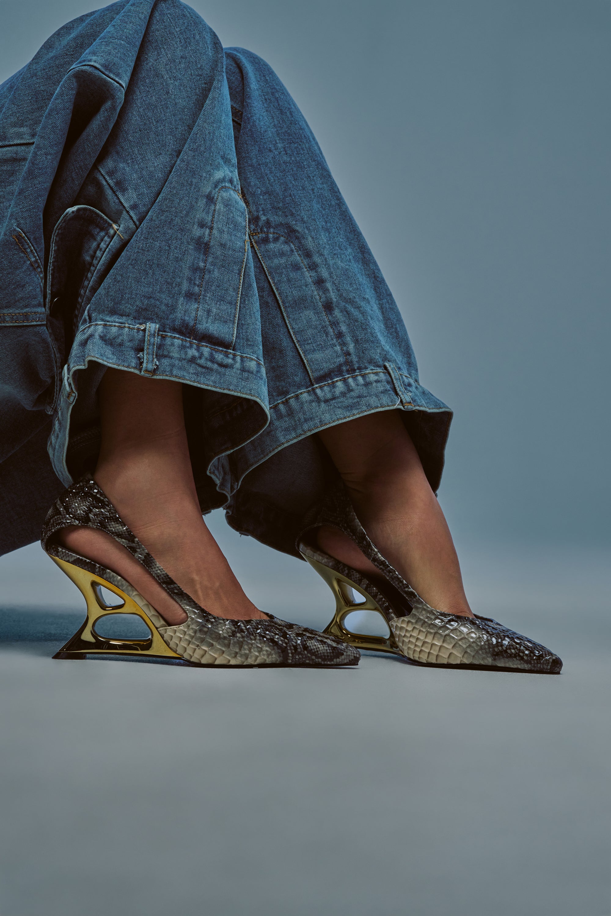 A person wearing wide-legged blue jeans and the AKIRA GOLD ABYSSICA SNAKE PRINT GOLD WEDGE HEEL PUMP, featuring unique open heels, sculptural wedge heels, and gold and black snake print accents, stands against a plain background.