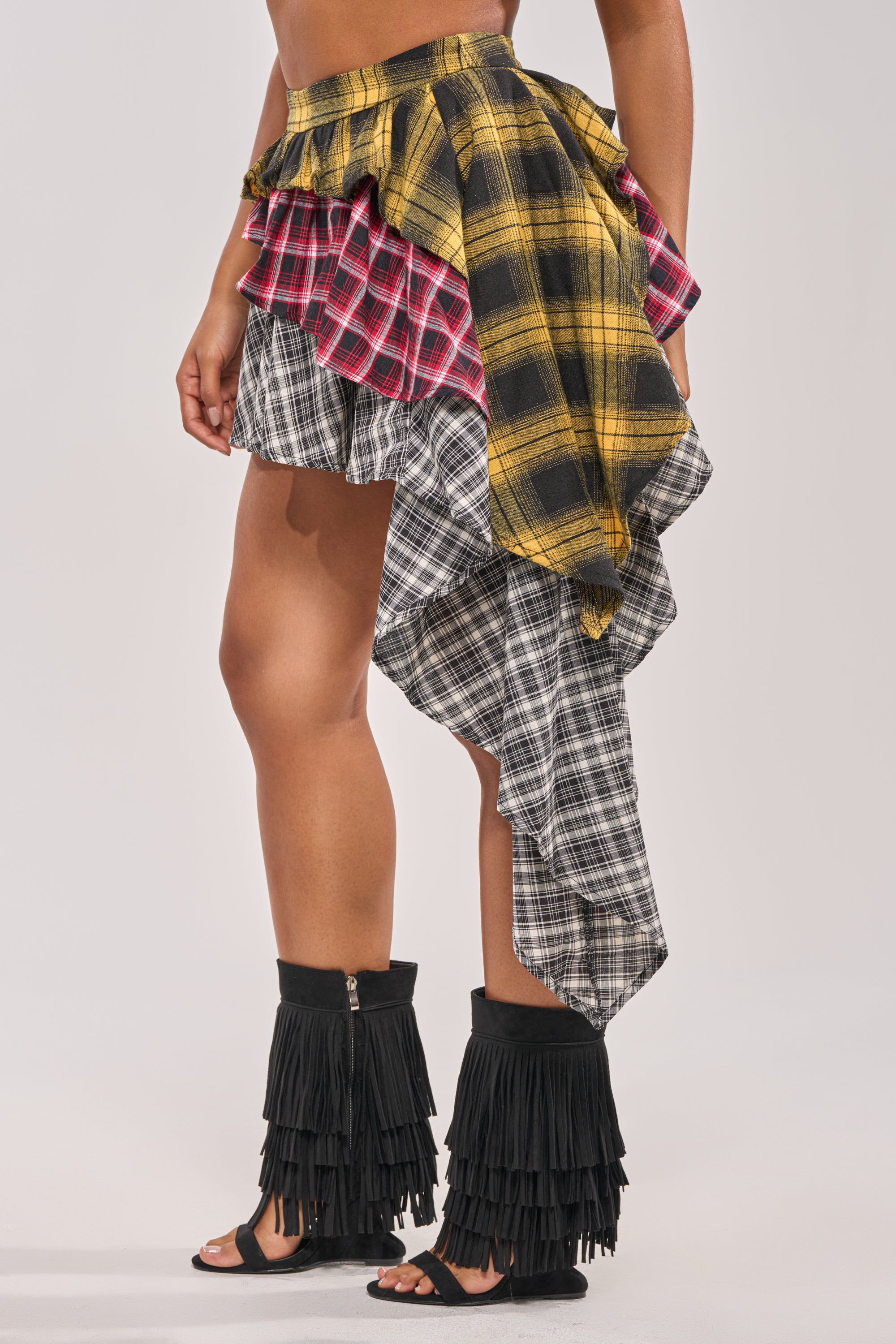 A person wears the ALL THAT WE HAVE TO DO PLAID MINI SKIRT in yellow, red, and black with asymmetrical ruffles, paired with black open-toe fringed boots. The image is cropped to show the skirt from the waist down. 2