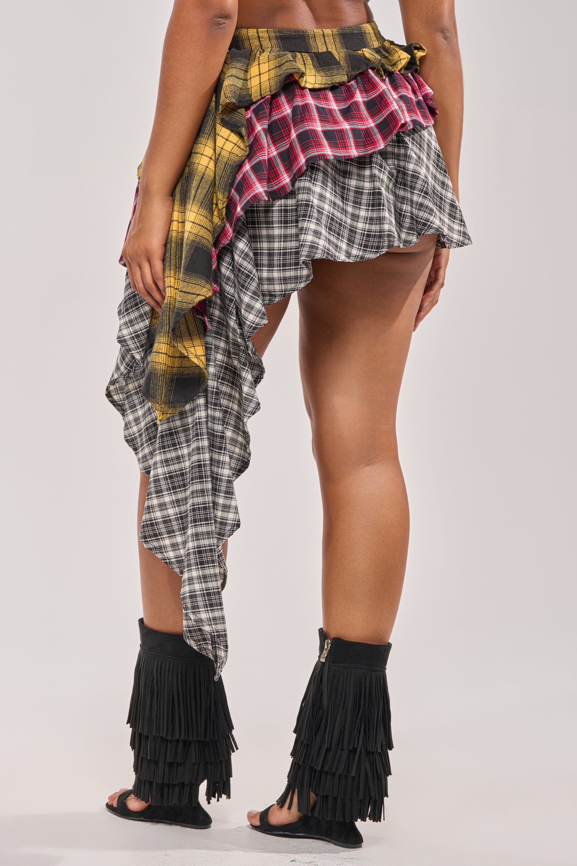 A person wearing the ALL THAT WE HAVE TO DO PLAID MINI SKIRT in yellow, red, and black, styled with black open-toe fringe sandals. The background is plain and light-colored. 4