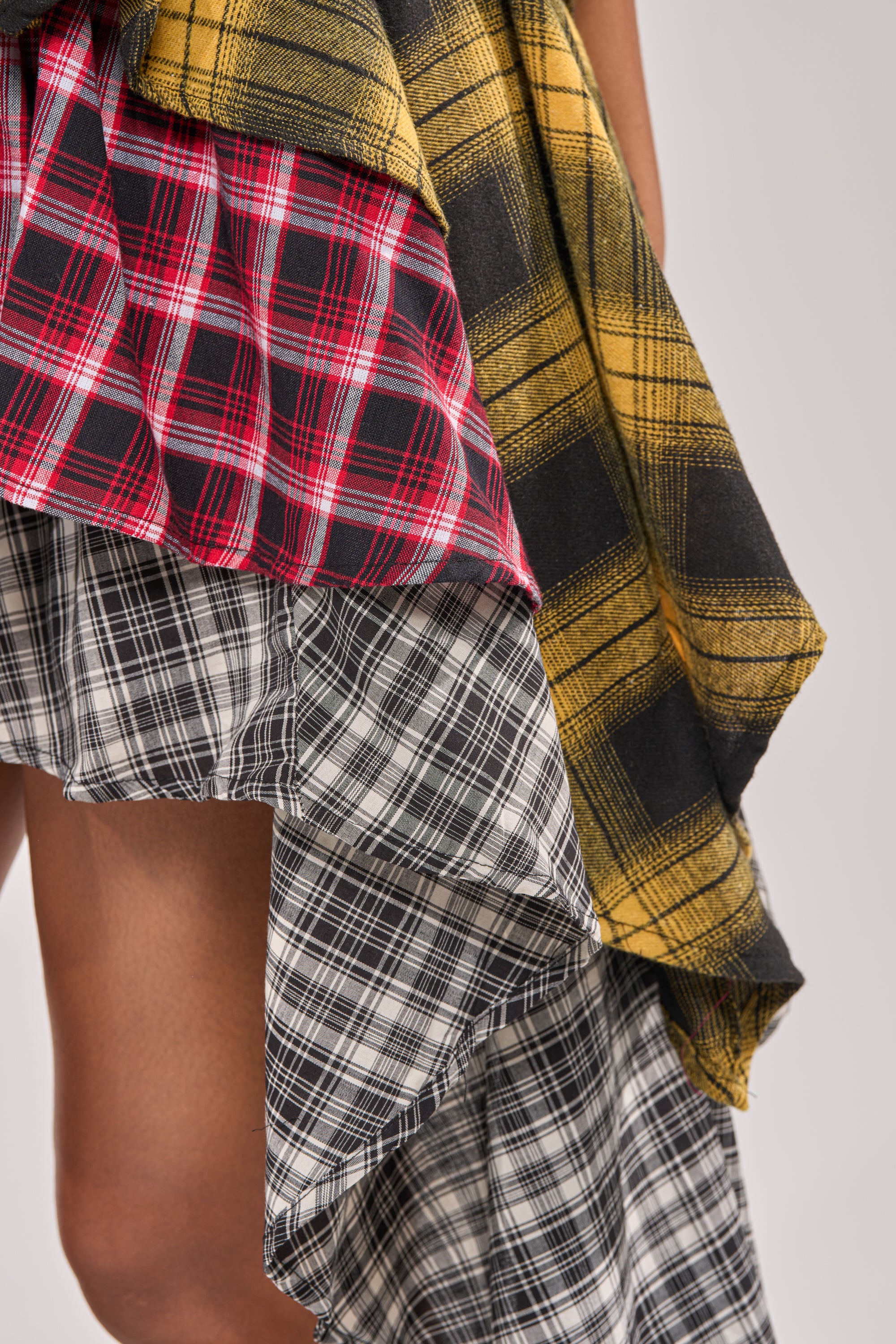 A close-up of a person wearing the ALL THAT WE HAVE TO DO PLAID MINI SKIRT, a festival streetwear-inspired ruffle mini made from mixed red, black, white, and yellow fabrics with asymmetrical overlapping panels. 6