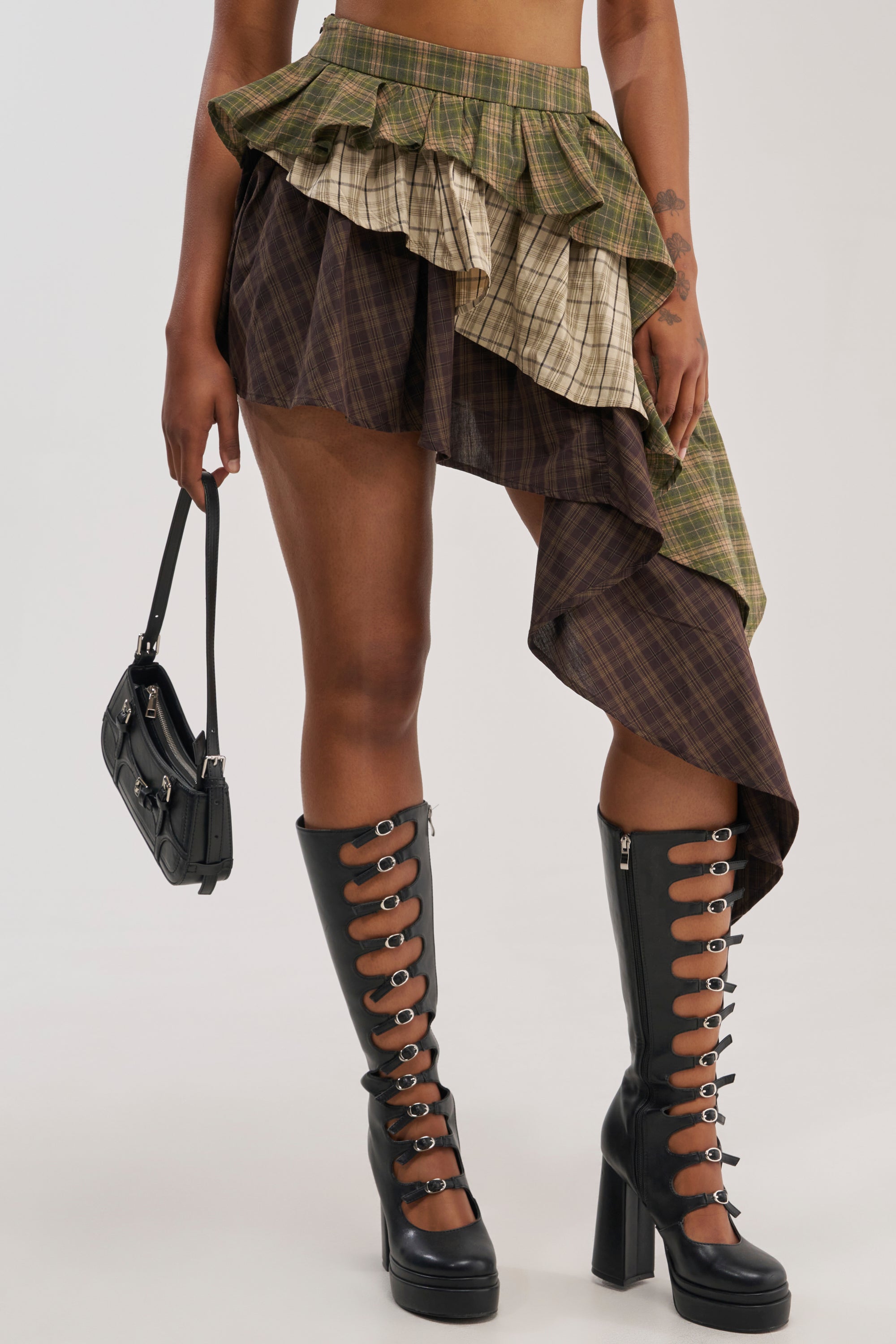 A person wears the SHAY MULTI PLAID MINI SKIRT, featuring layered, asymmetrical ruffles, paired with black lace-up knee-high boots with chunky heels and holds a small black handbag. The plain, light-colored background highlights this stylish festival streetwear look.