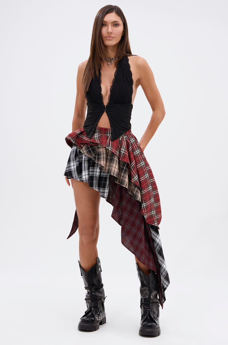 A woman with straight brown hair wears a black lace top and the SHAY MULTI PLAID MINI SKIRT—an asymmetric mini in red, black, and beige plaid—styled with black buckled boots, embracing grunge fashion against a plain white background.