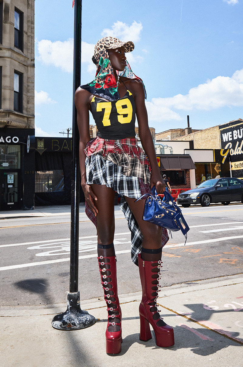 A person stands on a city sidewalk wearing a sporty black tank, tall red platform boots, a cheetah-print cap, floral scarf, red belt bag, blue handbag, and the SHAY MULTI PLAID MINI SKIRT.