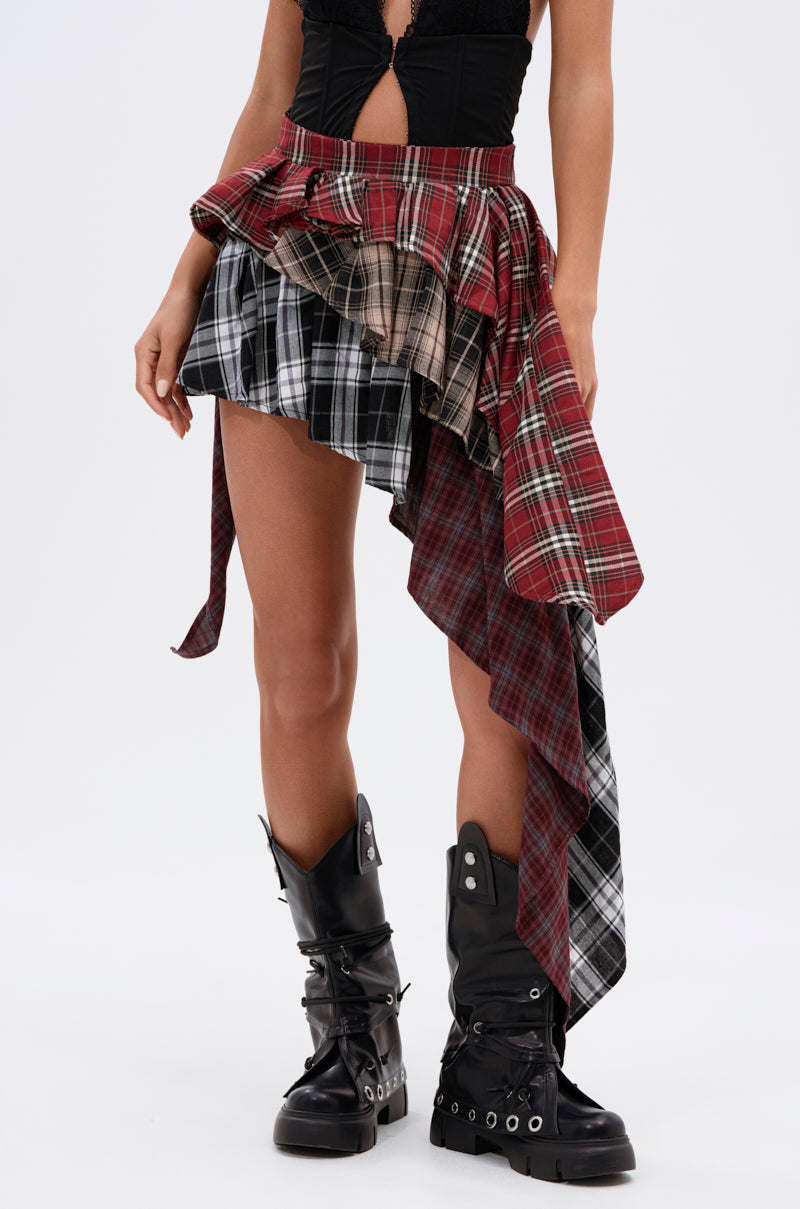 Wearing a black top, the person pairs it with the SHAY MULTI PLAID MINI SKIRT—an asymmetric high-low mini featuring layered red and black tartan—and black buckle boots, standing against a white background for bold grunge style.