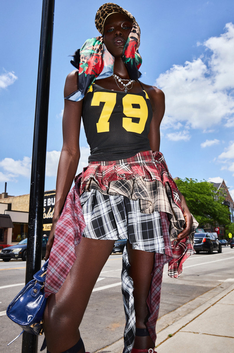A person stands on a city sidewalk wearing the SHAY MULTI PLAID MINI SKIRT, a black tank top with yellow "79," a red plaid shirt tied at the waist, a colorful scarf, and holding a blue handbag under a bright sky.