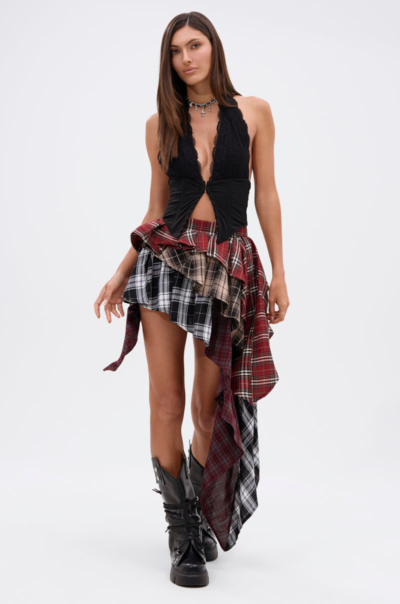 A woman models the SHAY MULTI PLAID MINI SKIRT with an asymmetric hemline, styled with a black lace top and tall boots. Her long brown hair and neutral expression enhance the grunge vibe against a plain white backdrop.