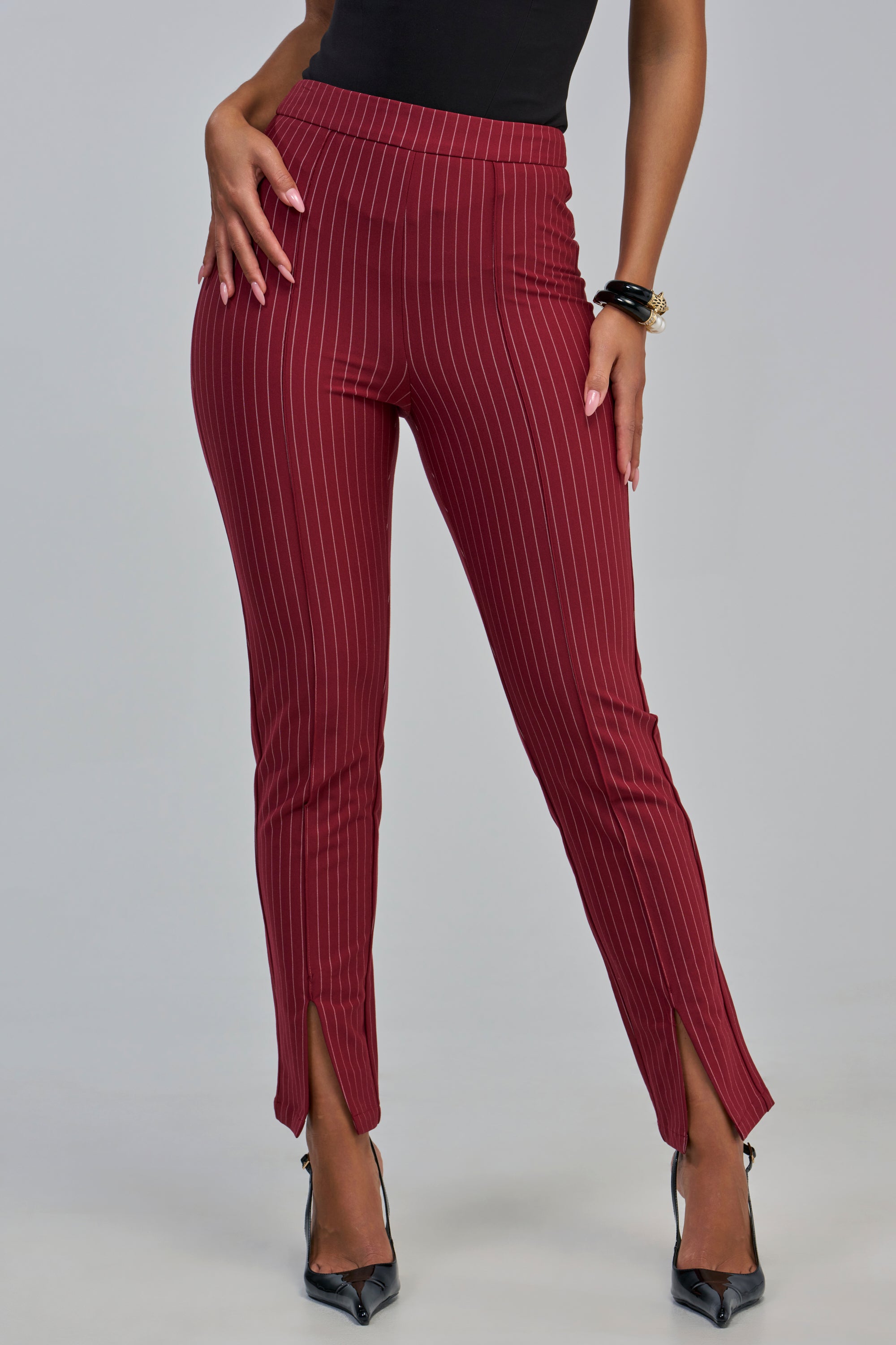 A woman models the LIVI Stretchy Pinstripe Skinny Legging with Slits in Burgundy, styled with a black top, black pointed heels, and bracelets. Featuring a high-rise fit and front ankle slits, these pants offer chic workwear appeal as she poses hands-on-hips against a gray background.