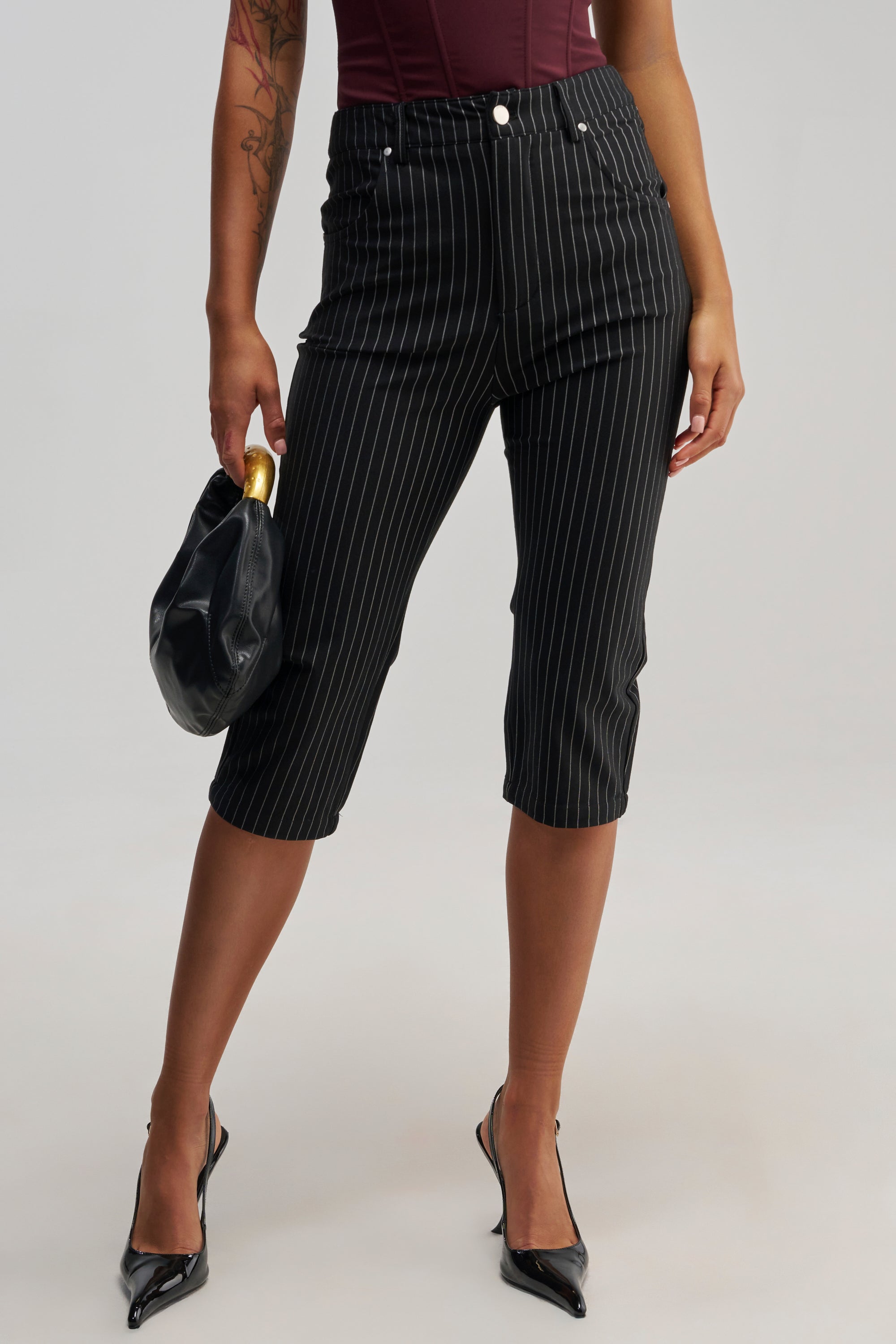 A person models modern business casual style, wearing the BUSINESS ONLY CAPRI—black pinstripe capri pants—paired with a burgundy top, black pointed heels, and a small black bag with a gold handle. Only the lower half of their body is shown, highlighting chic office wear.