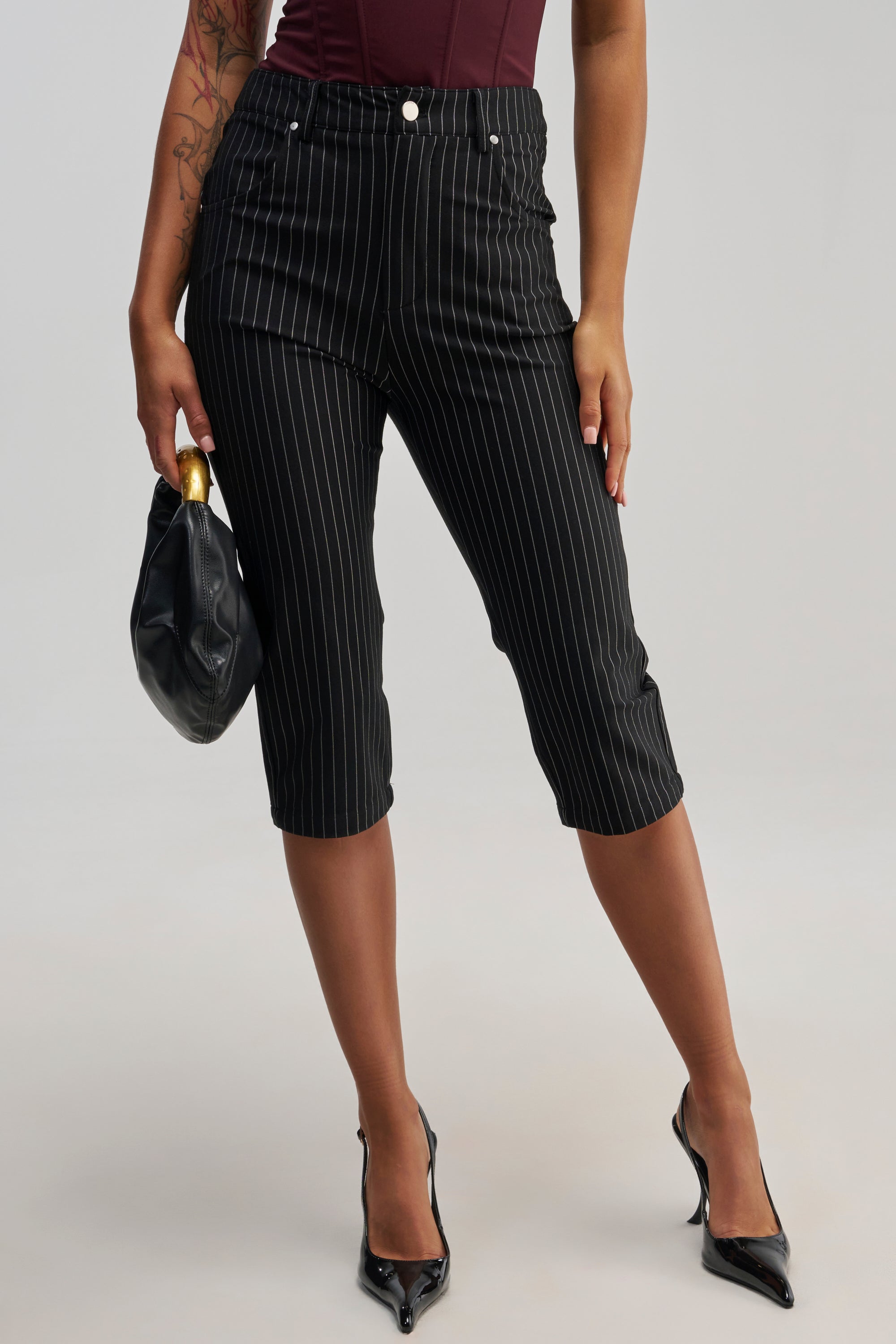 A person is seen wearing the BUSINESS ONLY CAPRI in black pinstripe, paired with black pointed heels and carrying a black handbag featuring a gold handle. The polished business casual ensemble is partially visible from the waist up, set against a plain, light background.