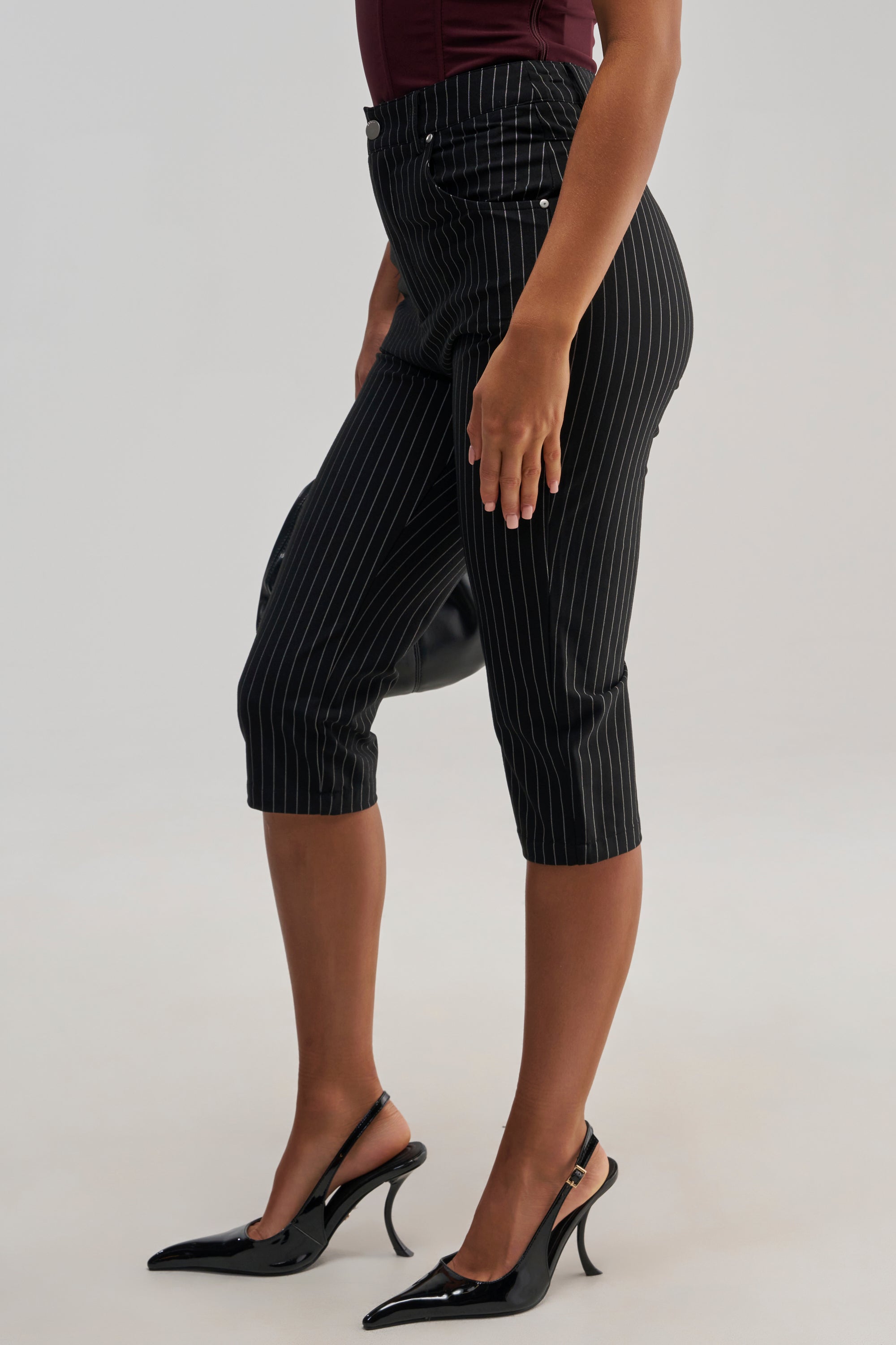Wearing the BUSINESS ONLY CAPRI in black pinstripe and paired with black pointed-toe slingback heels, this person stands against a plain background, effortlessly showcasing a chic business casual outfit that’s ideal for the office.