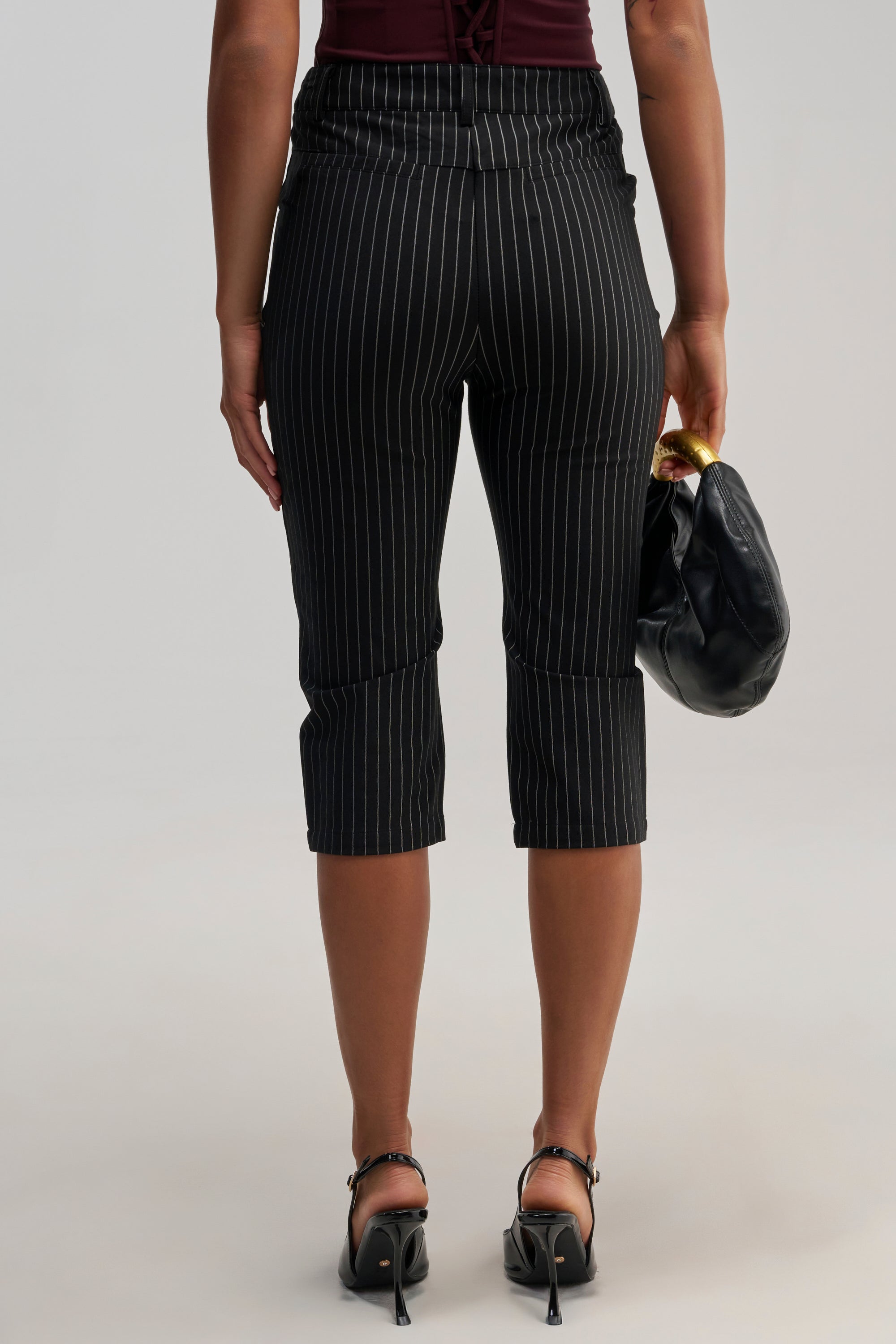 A person is seen from behind wearing the BUSINESS ONLY CAPRI in black pinstripes, paired with black heeled sandals and holding a black clutch bag featuring a gold accent. The background is plain and light-colored.
