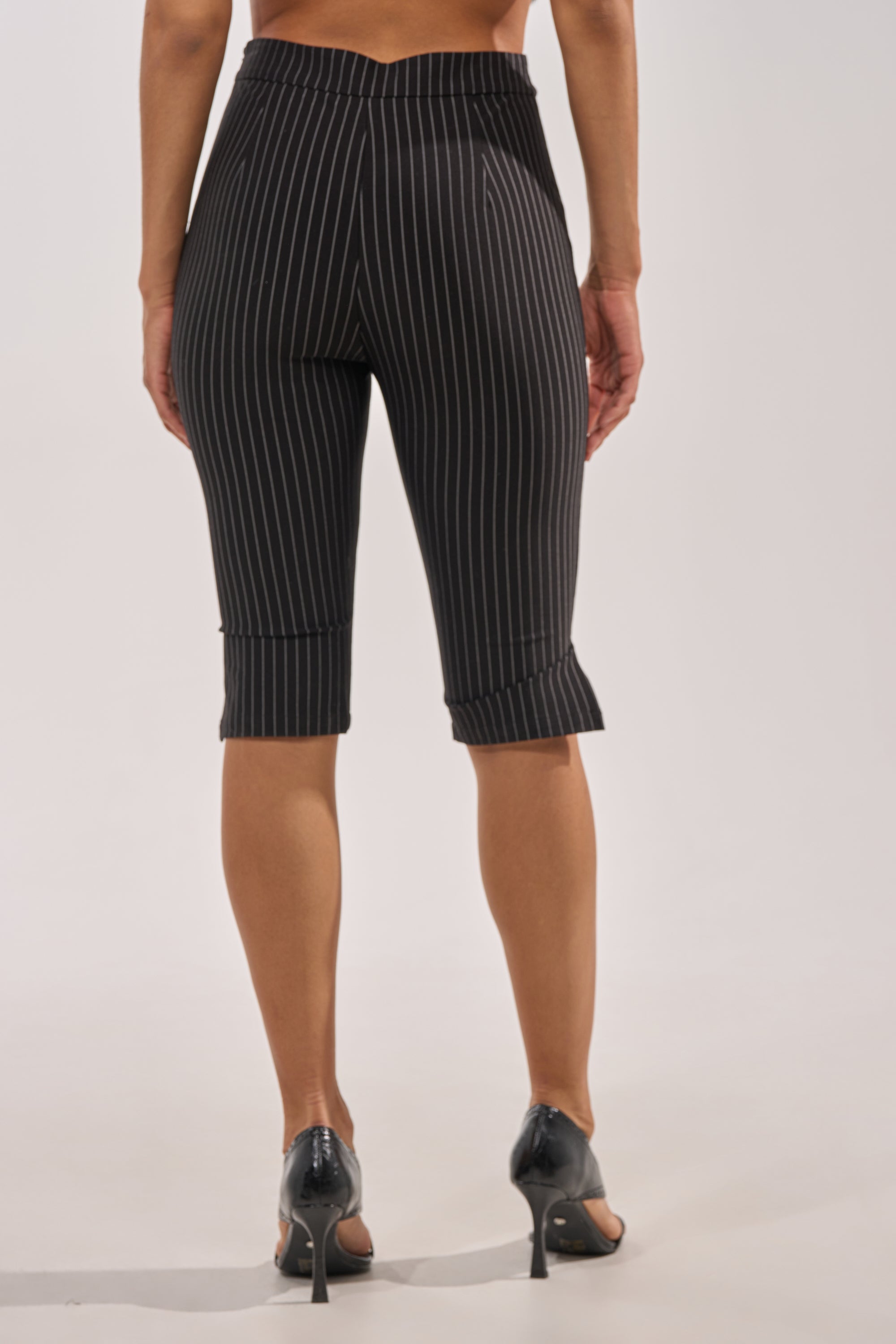 A person faces away, wearing BUSINESS ONLY CAPRI black pinstripe high-rise waist trousers and black high-heeled shoes against a plain light background. 4
