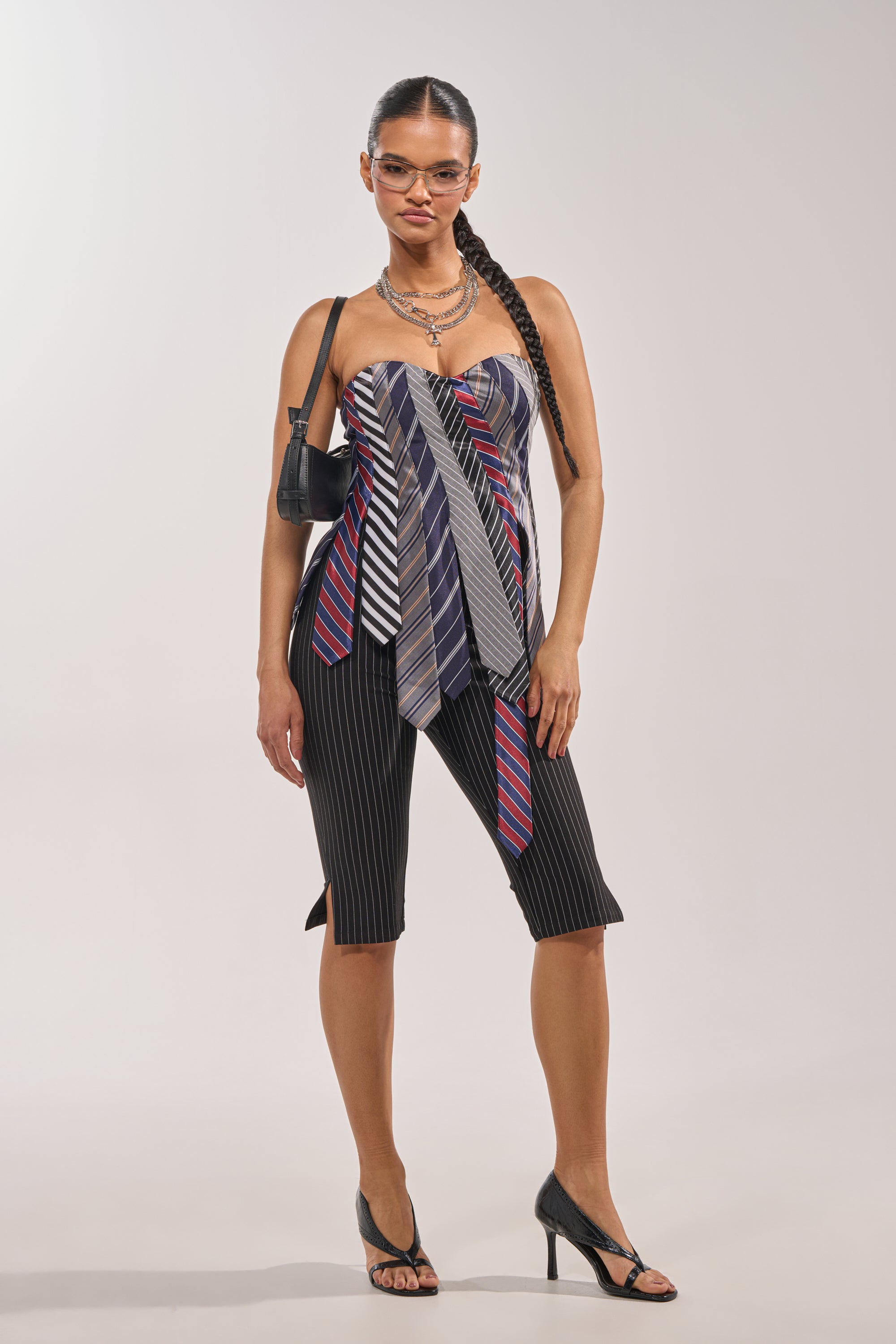 A woman stands confidently in the BUSINESS ONLY CAPRI, a business-casual pinstripe high-rise capri trouser. She pairs it with a strapless top of assorted neckties, layered necklaces, glasses, a small black shoulder bag, and open-toe heels. 5