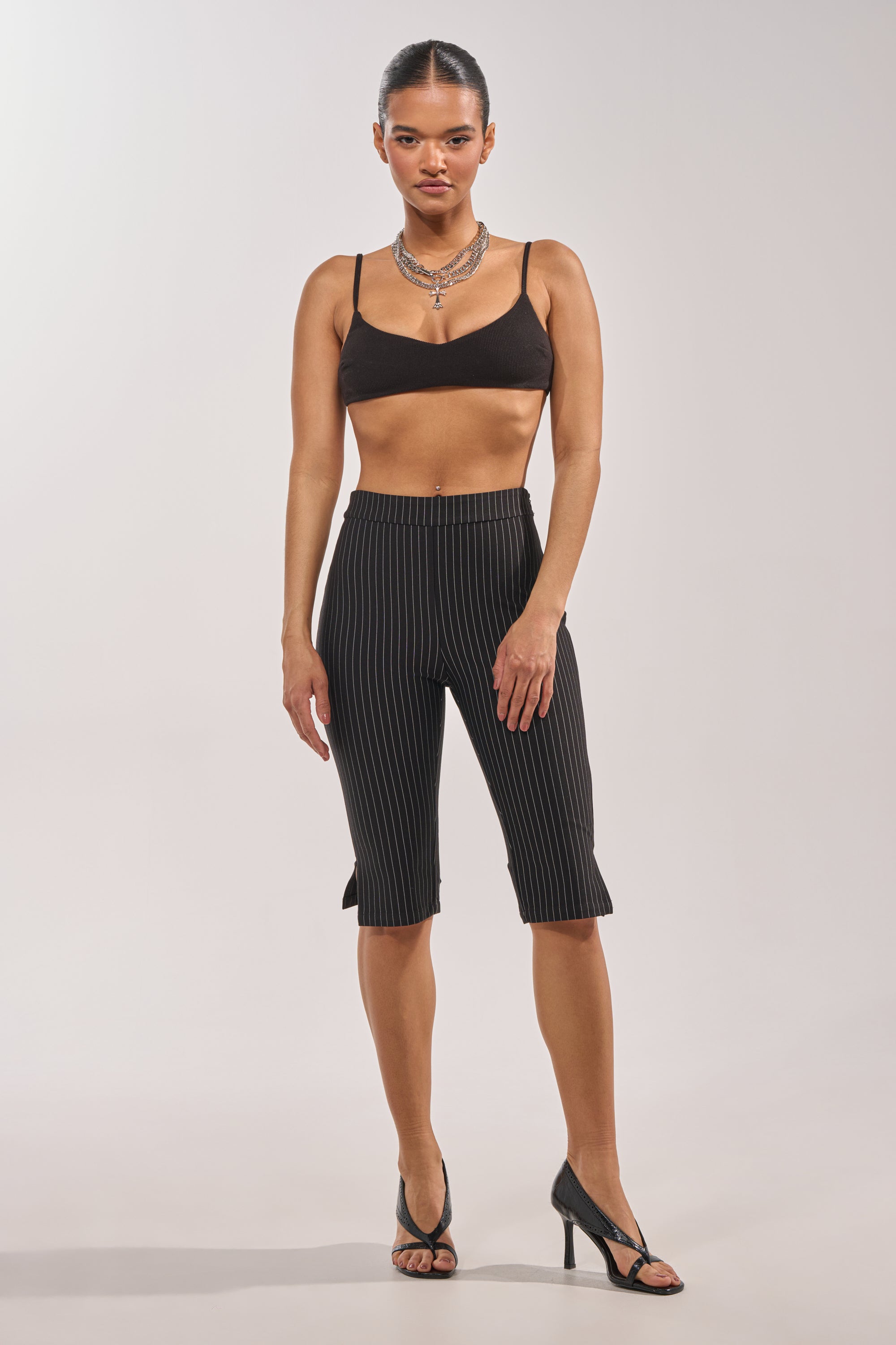A woman faces forward wearing the BUSINESS ONLY CAPRI high-rise slim-leg pinstripe capri trousers, a black crop top, black high heels, and a silver necklace against a plain light background. 6
