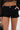 Wearing the BUSINESS CASUAL PINSTRIPE SHORT—high-rise, stretchy black pinstripe shorts with a gold button—paired with a black lace top and a chic black handbag featuring a gold handle. One hand rests casually in the pocket.