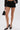 A person wears BUSINESS CASUAL PINSTRIPE SHORTS with a high-rise fit, a black lace long-sleeve top, black pointed-toe heels, and holds a small black handbag with a gold handle against a plain white background.