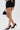 A person wears the BUSINESS CASUAL PINSTRIPE SHORT in a high-rise fit, a sheer long-sleeve lace top, black pointed-toe heels, and holds a black handbag with a gold handle. The background is plain white.