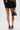 A person stands with their back to the camera, wearing the BUSINESS CASUAL PINSTRIPE SHORT in black, paired with a sheer lace top, black high heels, and holding a black handbag with a gold handle.