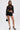 A woman poses against a white background in the BUSINESS CASUAL PINSTRIPE SHORT, paired with a sheer black lace long-sleeve top, a black bralette, black high heels, and a small black handbag.