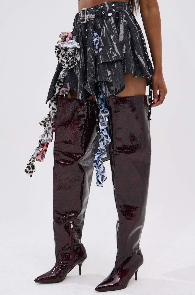 A person wearing glossy, dark maroon thigh-high boots with pointed toes and kitten heels, paired with the FEELING WILD MINI SKIRT featuring a high-waisted asymmetrical cut with straps and colorful animal print fabric strips.