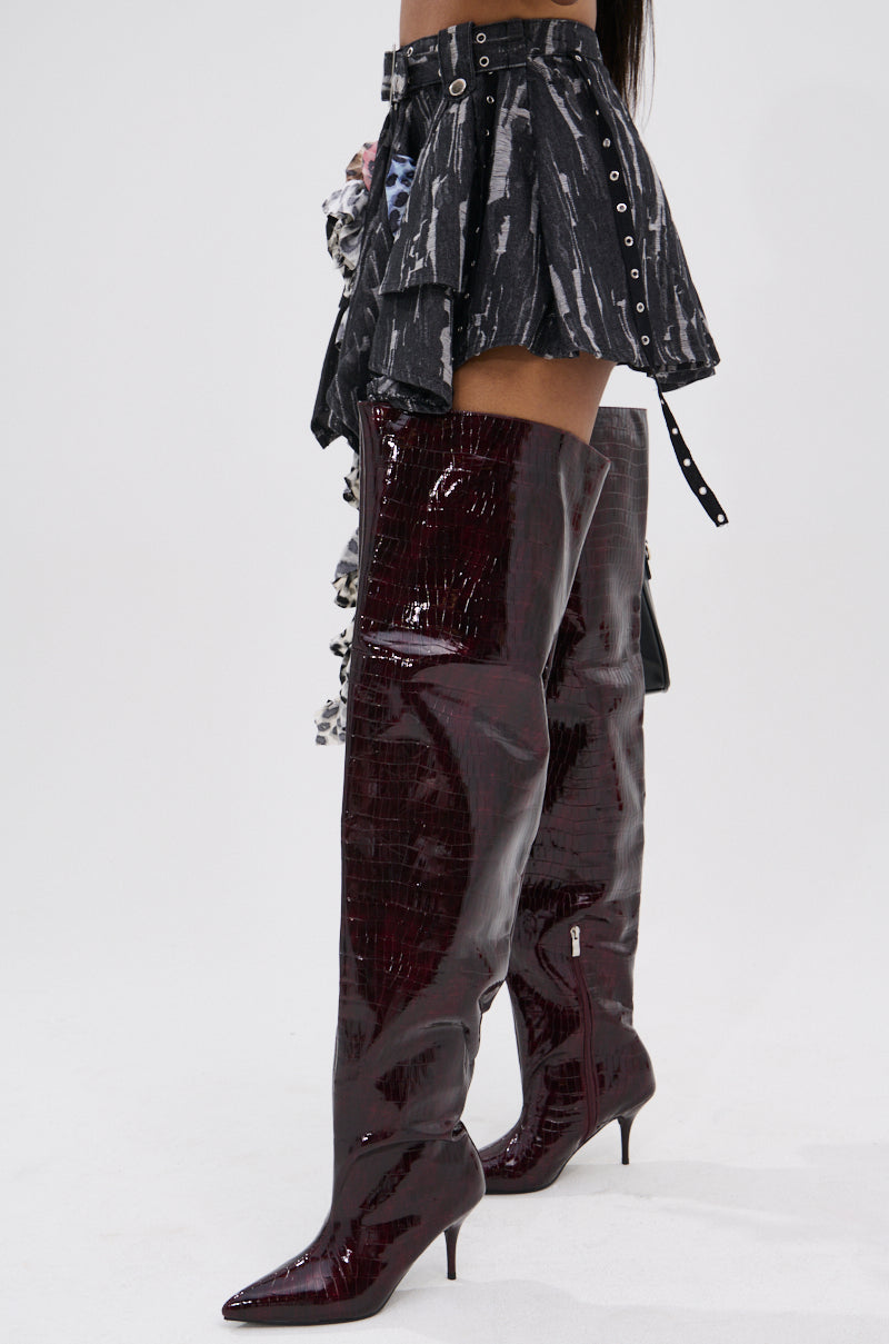 Wearing the FEELING WILD MINI SKIRT—short, asymmetrical, black with belt details—and shiny dark burgundy knee-high pointed boots, a person stands against a plain white background.
