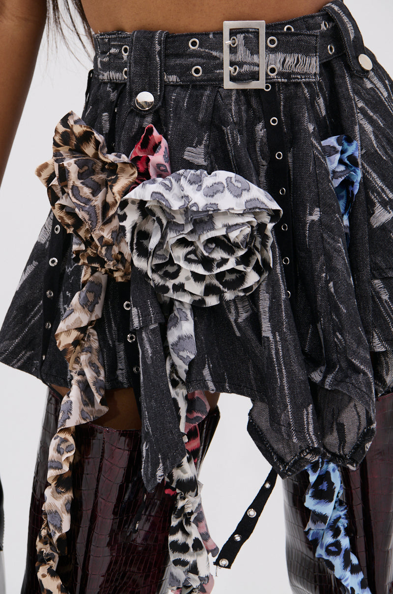 A close-up of the FEELING WILD MINI SKIRT, an asymmetrical black denim mini with a silver belt buckle and bold animal print fabric rosettes; paired with shiny textured dark red boots.