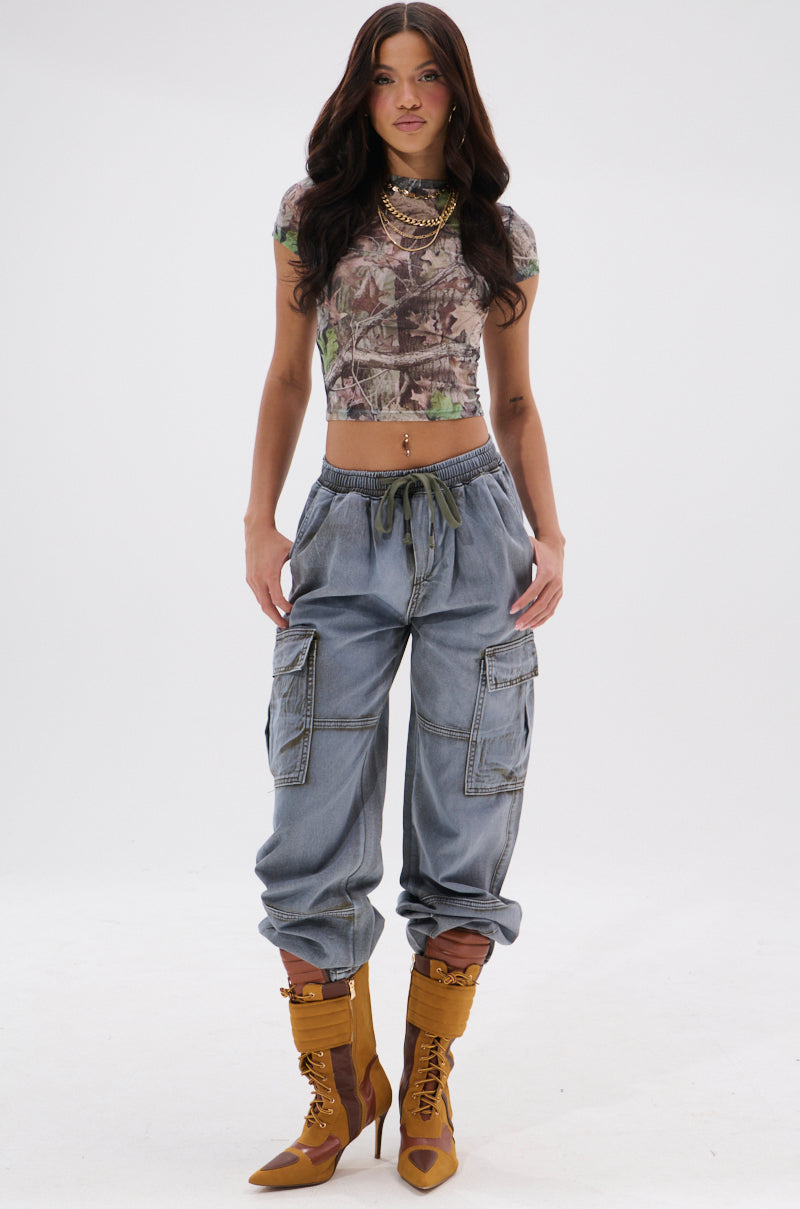 A woman faces forward against a white background, wearing the WALLEY CARGO JOGGER pants with pockets, a cropped camo top, tan lace-up heeled boots, and layered gold necklaces. 0