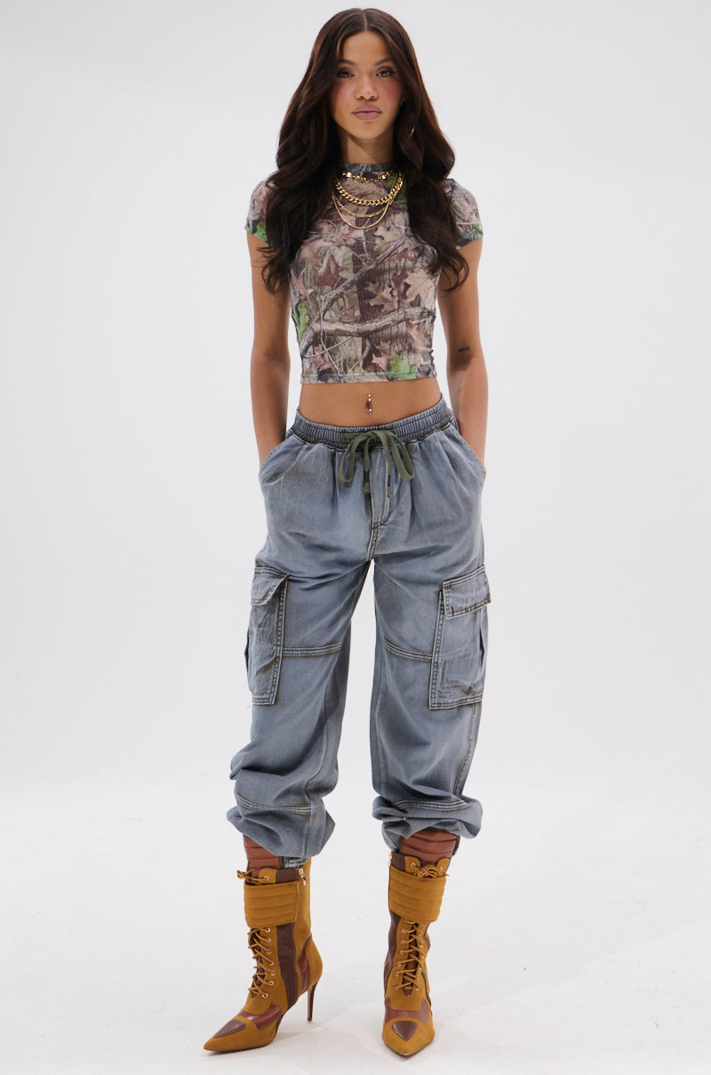 A woman with long wavy hair and layered gold necklaces poses in front of a white background wearing the WALLEY CARGO JOGGER, a fitted patterned crop top, and brown high-heeled lace-up boots. 6
