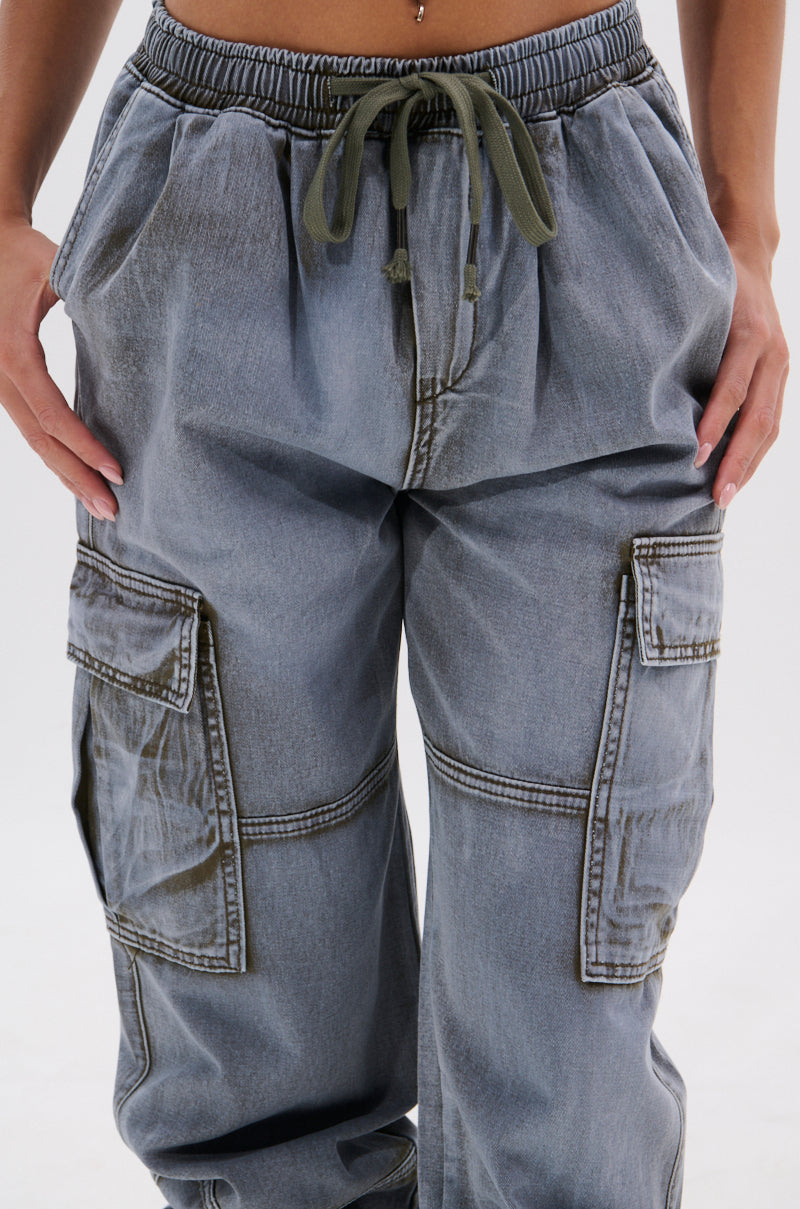 A person wears the WALLEY CARGO JOGGER, featuring loose-fitting, faded gray fabric, an oversized cargo pocket, and an olive drawstring waistband while standing with hands by their sides. 7