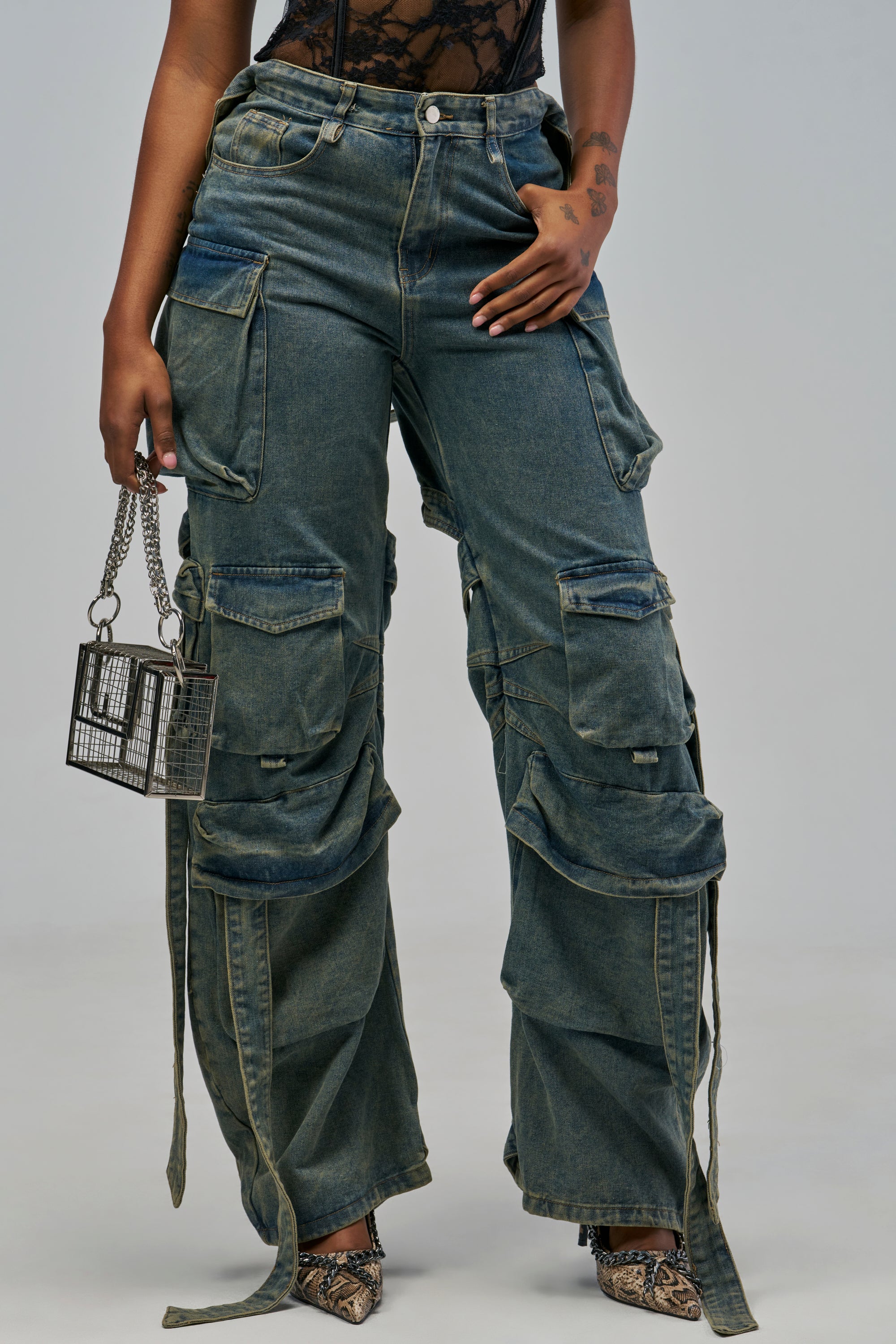 CHECK OUT THE BACK CUTOUT RELAXED DENIM: relaxed-fit wide-leg blue jeans featuring multiple pockets and long strap details, styled with a small metallic cage handbag and snakeskin-patterned heels.