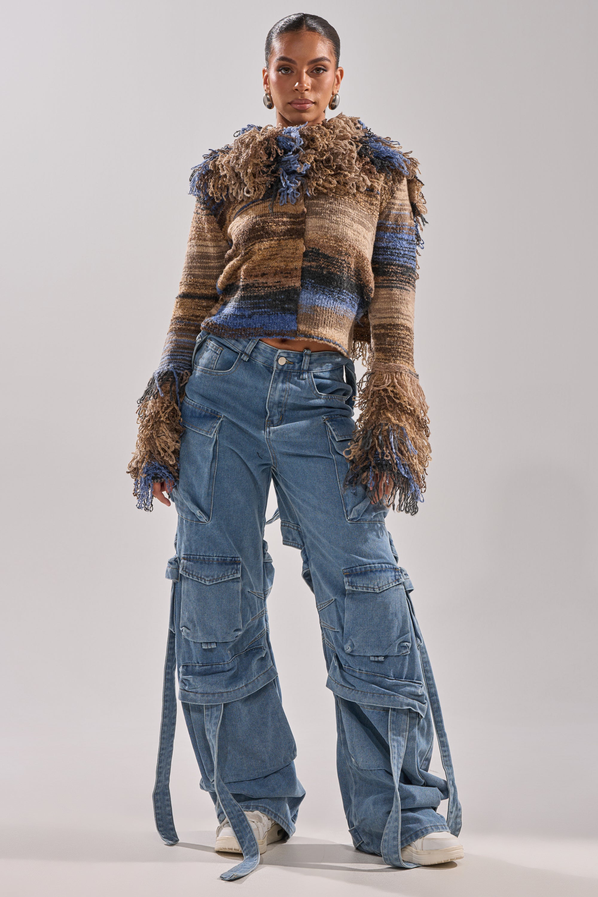 A woman stands confidently in a textured brown and blue striped sweater, wide-leg HAILEY WIDE LEG DENIM cargo pants with high-rise styling, large pockets, long straps, and white shoes against a plain background.