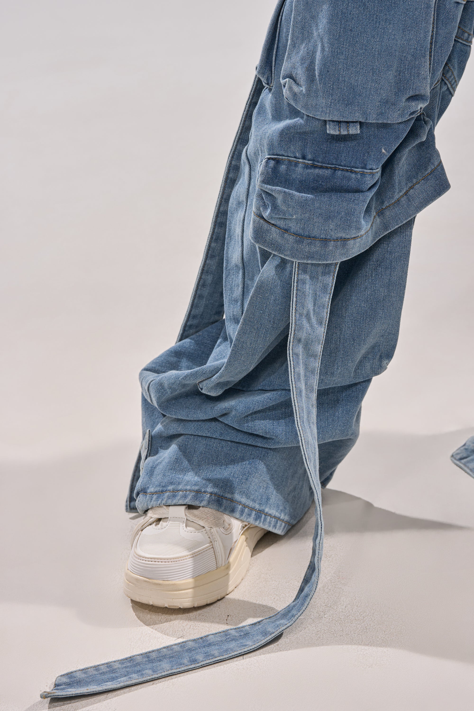 Someone wearing HAILEY WIDE LEG DENIM jeans with long, loose straps and cargo pockets pairs them with white platform sneakers while standing on a smooth, light surface.