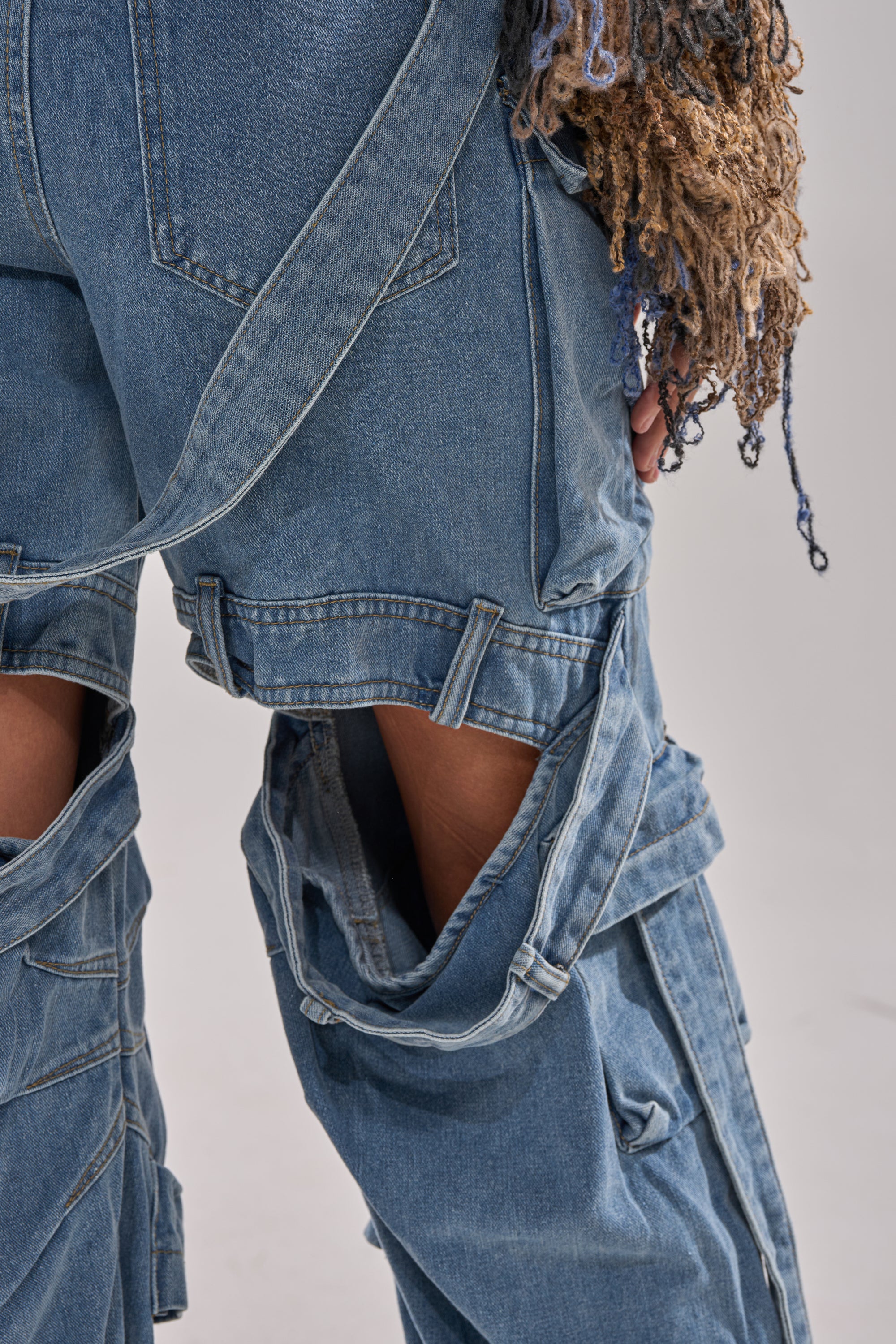 A close-up shows a person wearing HAILEY WIDE LEG DENIM—high-rise jeans with large cutouts, straps, and frayed multicolored yarn details. The wide-leg style partially covers their hand against a plain light background.