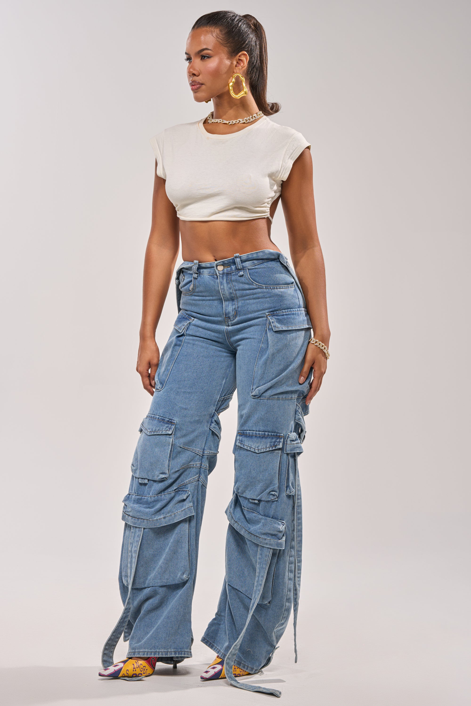 A woman poses in the CHECK OUT THE BACK CUTOUT RELAXED DENIM—blue wide leg cargo jeans with multiple pockets and straps—styled with a white crop top, colorful heels, gold hoops, a chain necklace, and her hair in a high ponytail. 0