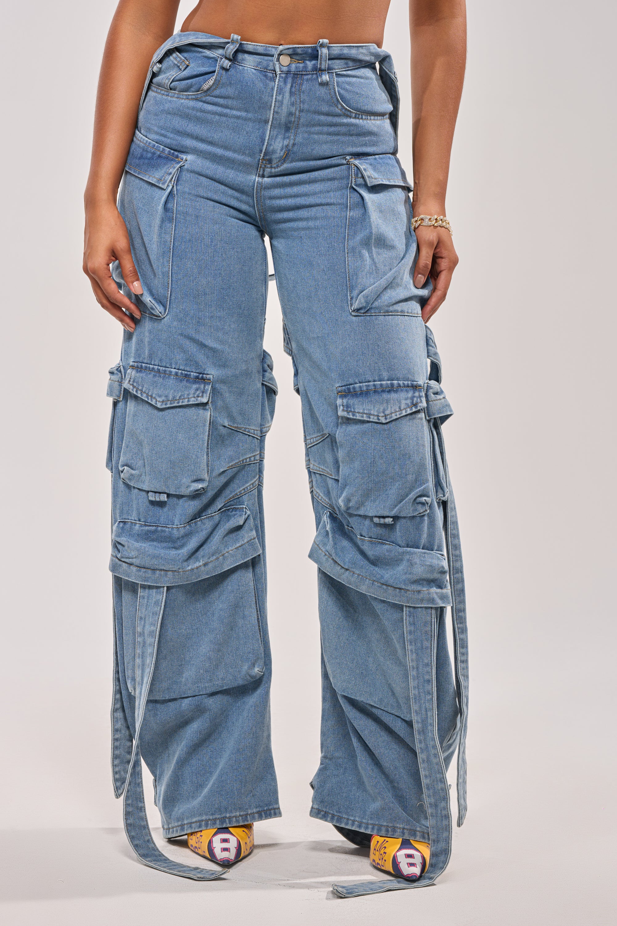 A person wearing CHECK OUT THE BACK CUTOUT RELAXED DENIM with multiple large pockets, buckles, and straps, styled with colorful socks and a gold bracelet, shown from waist to feet against a plain background. 1