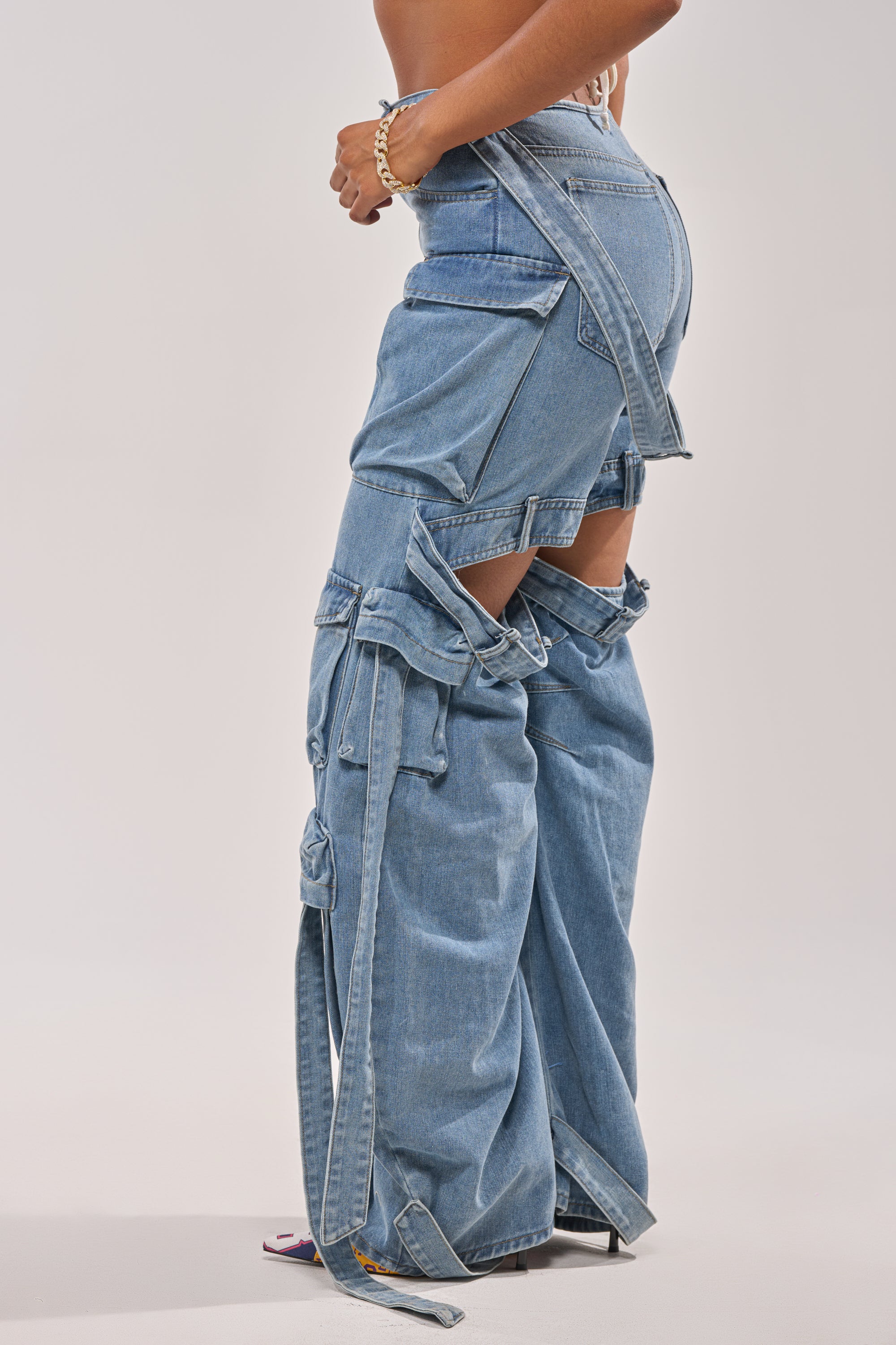 Someone wears the CHECK OUT THE BACK CUTOUT RELAXED DENIM—unique, layered wide-leg jeans with multiple straps, cutouts revealing legs and shorts, and a loose deconstructed design inspired by utility cargo styles. 2