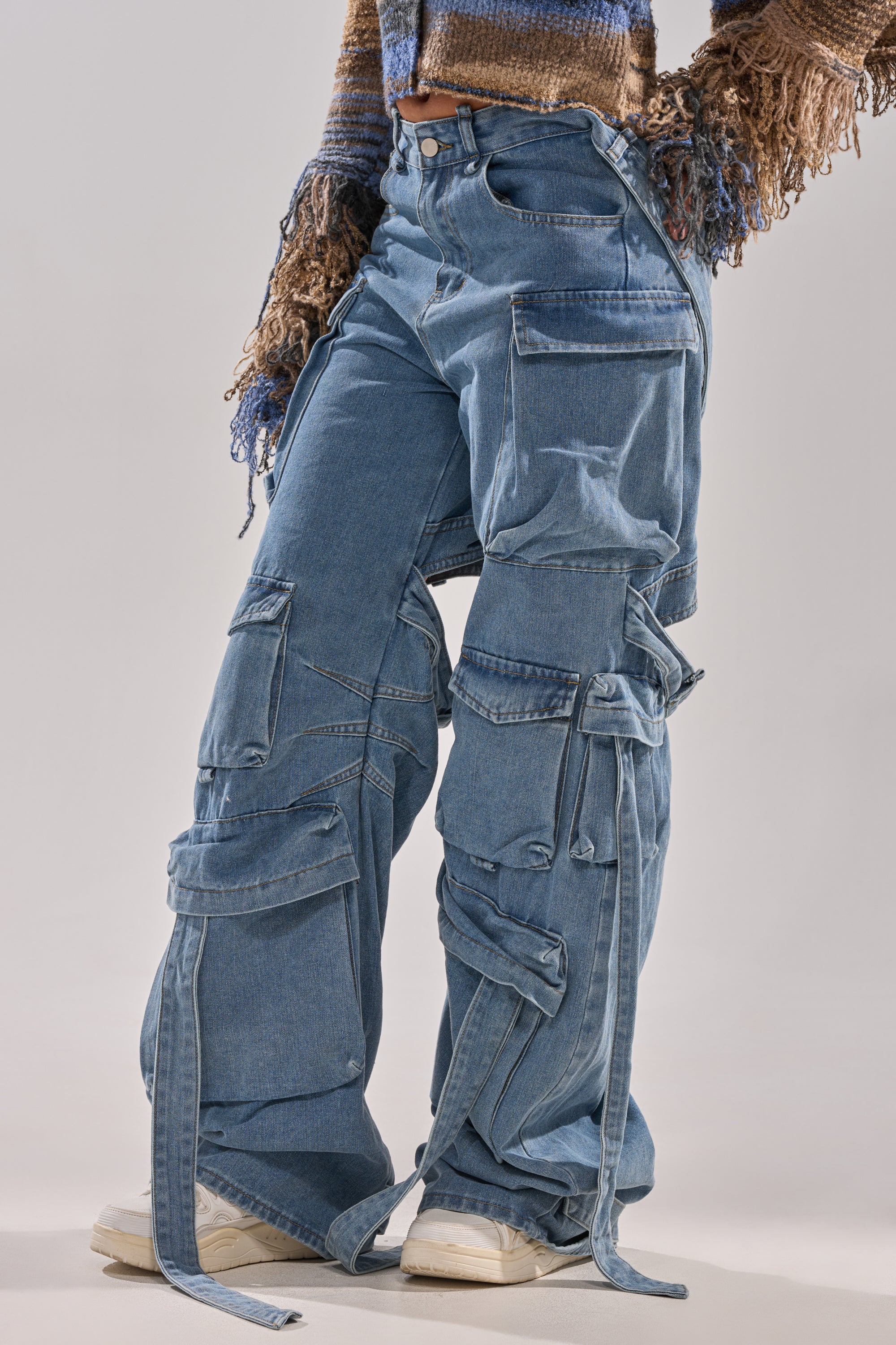 Someone is dressed in HAILEY WIDE LEG DENIM cargo jeans featuring multiple large pockets and straps, styled with a multicolored textured sweater and white sneakers. The person's face is not visible.