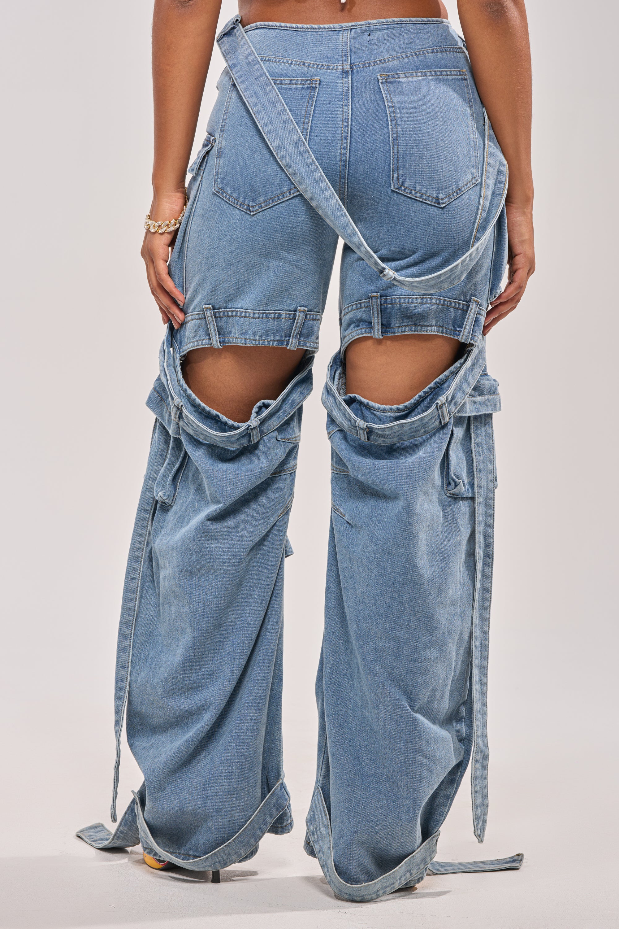 A person wearing CHECK OUT THE BACK CUTOUT RELAXED DENIM, featuring wide legs, large thigh cutouts at the back, visible pockets, and loose side straps. The image is shot from behind to show the lower back and legs. 3