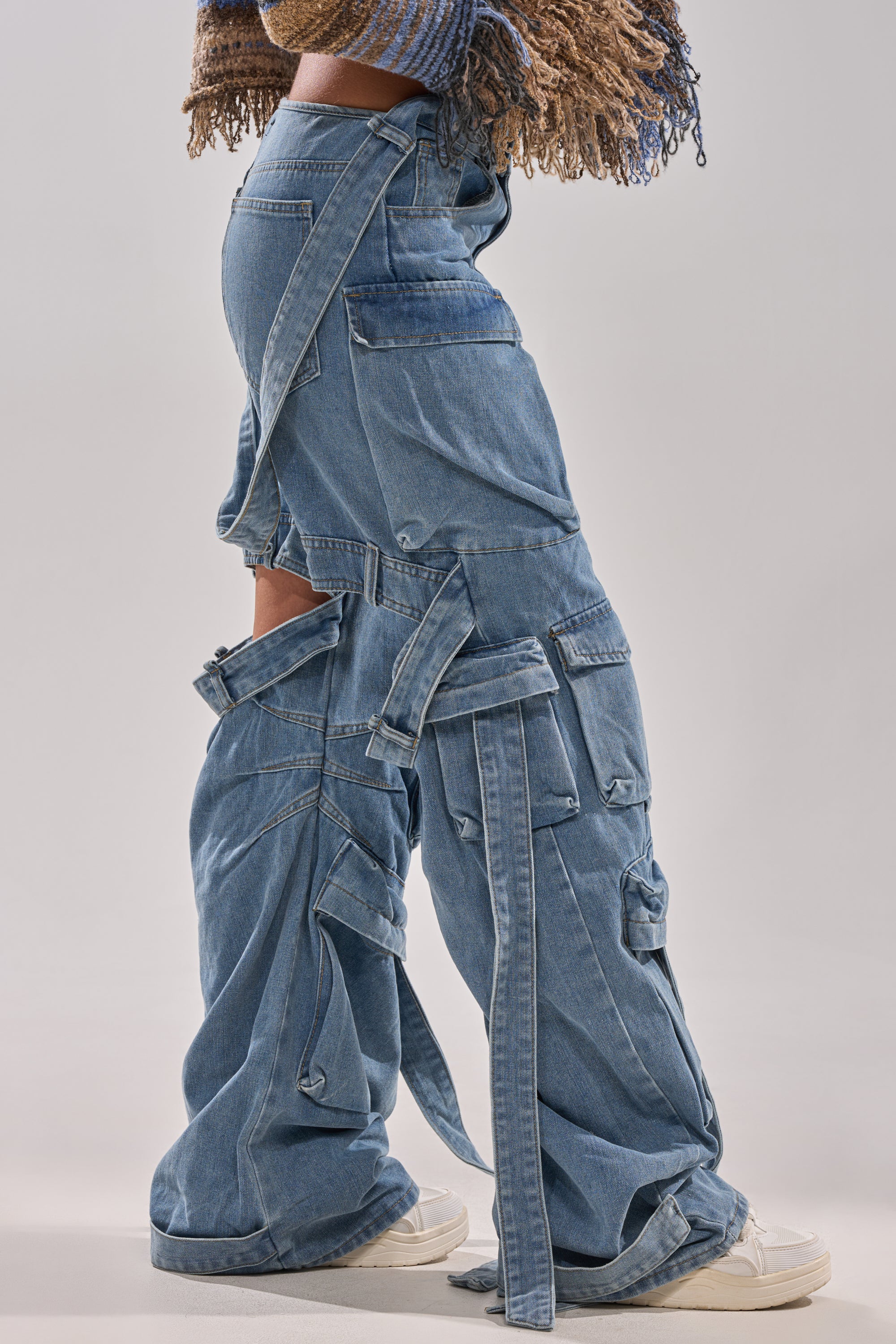 A person models the HAILEY WIDE LEG DENIM cargo jeans with multiple straps and panels, styled with white sneakers and a fringed sweater; only the lower half of their body is shown.