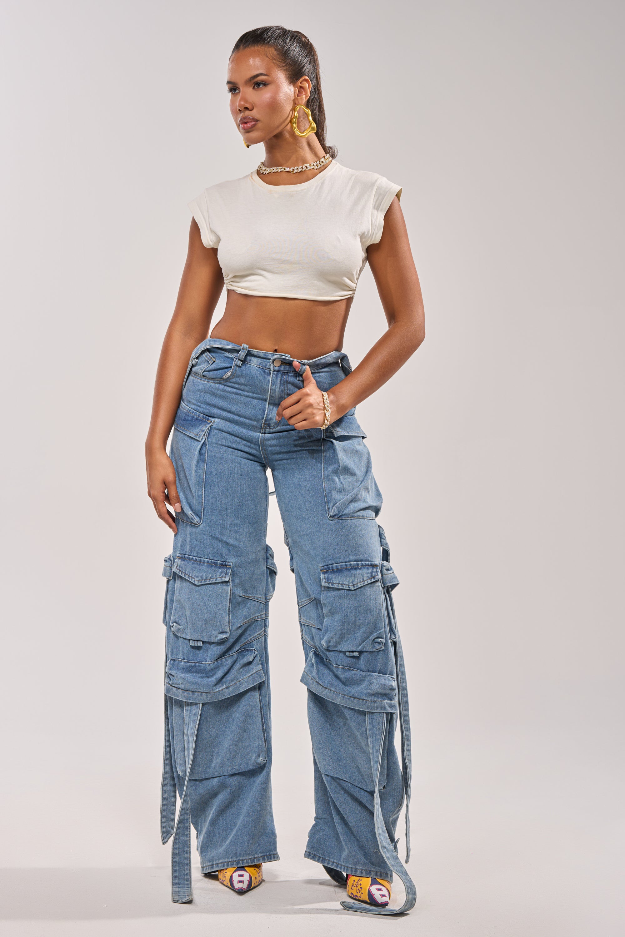 A woman stands confidently in a white crop top, chunky gold earrings, and the CHECK OUT THE BACK CUTOUT RELAXED DENIM cargo jeans with multiple pockets and straps, paired with open-toe heeled sandals against a plain light background. 4