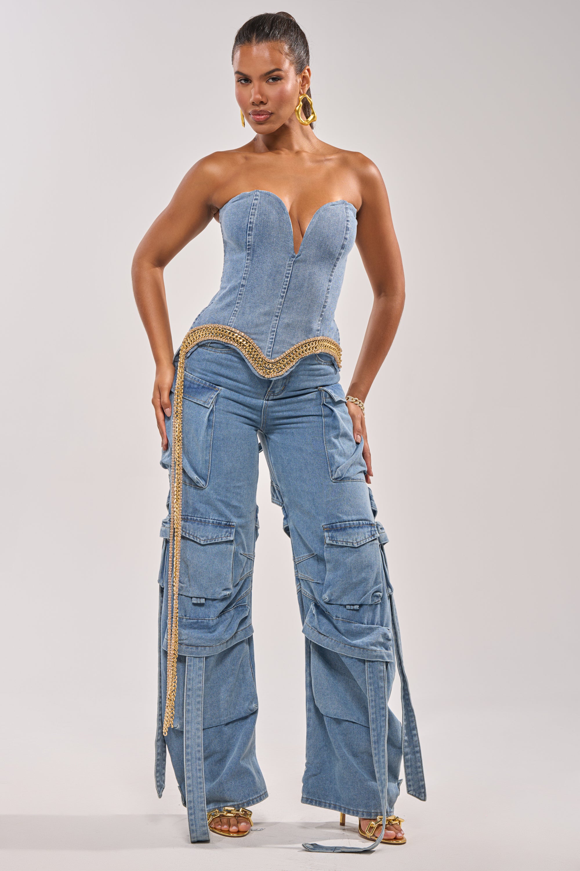 A woman models the CHECK OUT THE BACK CUTOUT RELAXED DENIM: a strapless, form-fitting bustier top with gold chain detail and matching high-rise, wide-leg cargo jeans featuring multiple pockets and straps. She accessorizes with gold hoops and heeled sandals. 6
