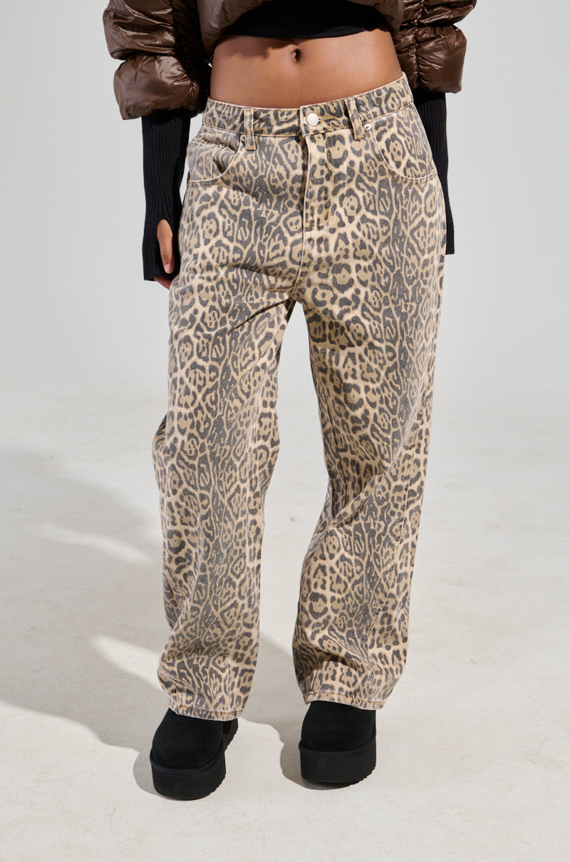 Wearing a pair of WILD CHILD LEOPARD PRINT DENIM JEANS, someone pairs them with a brown puffy jacket and completes the ensemble with black boots, their hands gently tucked into the jacket's sleeves. The backdrop is a plain, light gray. 0