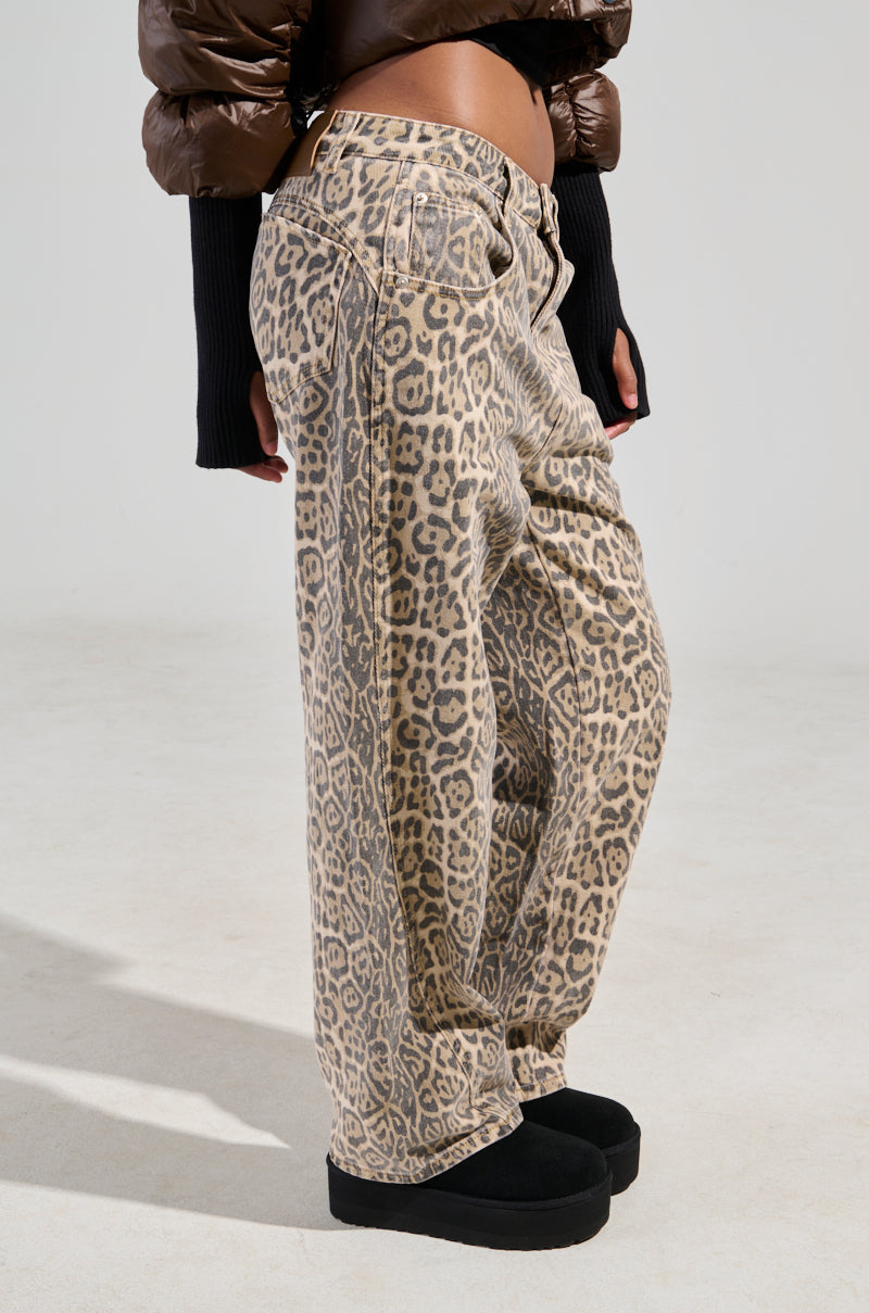 A person wearing WILD CHILD LEOPARD PRINT DENIM JEANS featuring a wide leg silhouette, paired with a cropped brown puffy jacket and black platform shoes, stands against a plain background. The jeans have a loose fit, and their hands are tucked into the pockets. 1