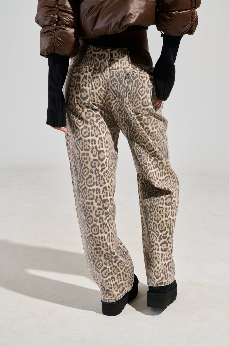 The individual is showcasing the WILD CHILD LEOPARD PRINT DENIM JEANS paired with a black top and a brown puffer jacket. Completing the look are black boots, all set against a simple, light backdrop. The image emphasizes the wide-leg silhouette and highlights the back view of the jeans. 3