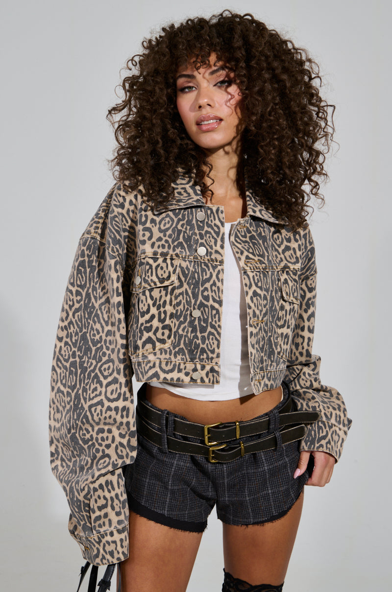 A person with curly hair is wearing the "NO CHEATING PRINTED DENIM JACKET," styled with its leopard print design over a white top, paired with plaid shorts and a double-belted waist. They stand against a plain background, exuding confidence with one hand on their hip in the chic cropped silhouette.